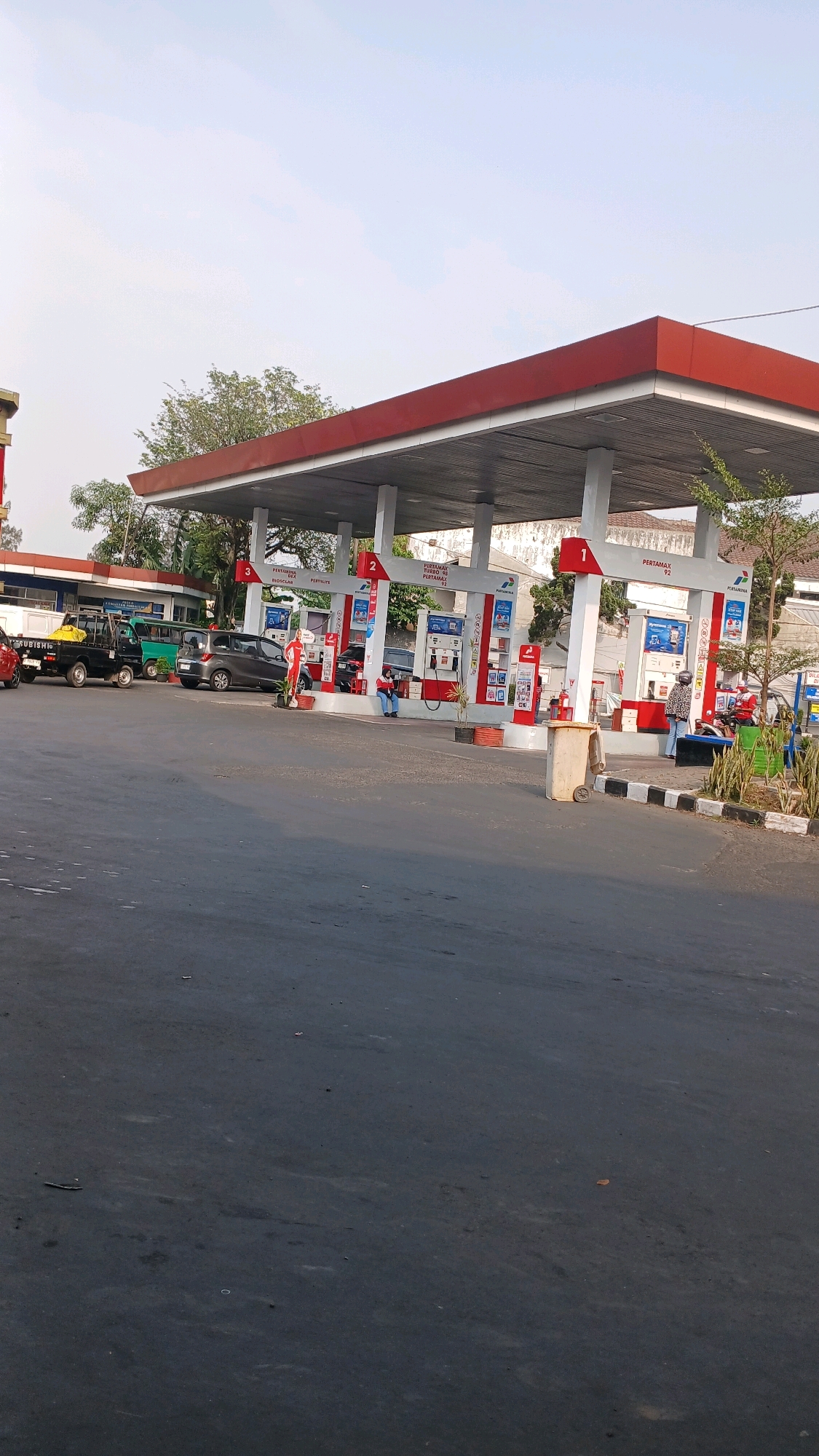 The image depicts a Pertamina gas station in Bandung City, Indonesia, with its distinctive red and white canopy. Several vehicles are visible, including a black pickup truck with a yellow object in its bed, a dark gray sedan, and other cars near the pumps. People are present at the gas station, with one individual seated near a pump and another standing. The scene shows the typical operations of a gas station, with pumps dispensing fuel. The environment is an outdoor setting under a clear sky, with trees and buildings in the background. The asphalt road in the foreground shows some wear and tear.