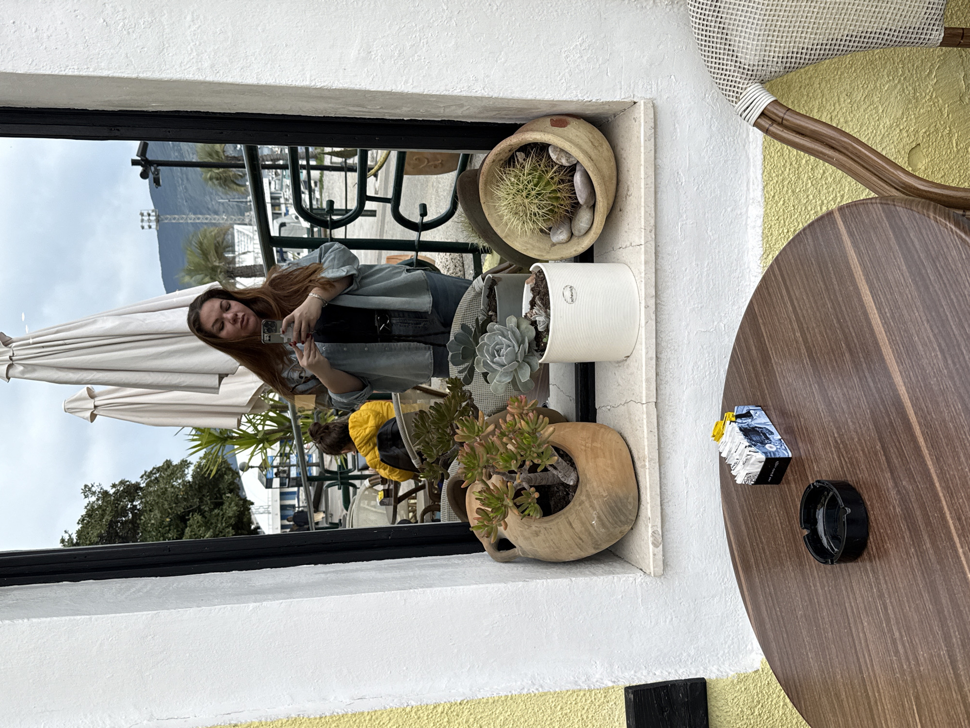 A person is taking a picture of themself in a mirror on a windowsill. The windowsill contains several potted plants, including cacti and succulents. The reflection shows a person in a jacket, an umbrella, other tables and chairs, and other patrons. The foreground includes a round wooden table with an ashtray and a pack of cigarettes on top, as well as the edge of a woven chair. Based on the location and architectural style, the image likely depicts an outdoor cafe scene in Herceg Novi, Montenegro.