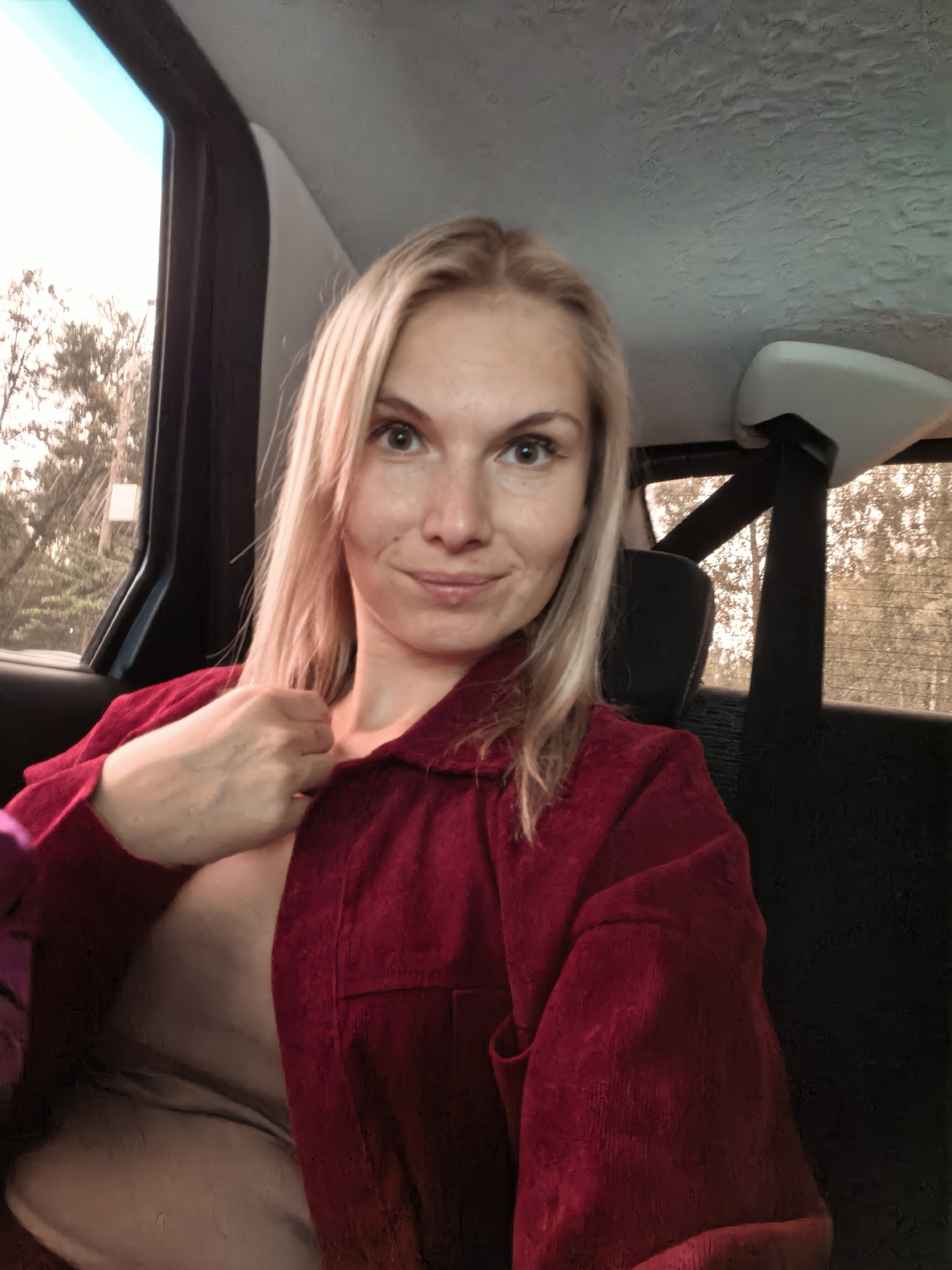 A young woman with blonde hair and green eyes is sitting in the back seat of a vehicle, looking at the camera. She is wearing a red corduroy jacket over a beige top. The jacket is partially unbuttoned, revealing her chest. The background shows trees and foliage through the car windows, suggesting an outdoor setting. The interior of the car is visible, including a headrest and a seatbelt. The overall lighting suggests it is daytime, possibly late afternoon given the warm tones. There is no specific indication of the city of Sterlitamak, Russia.