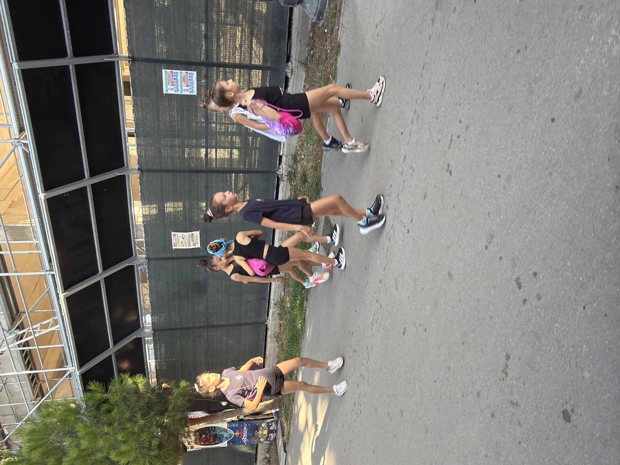 A group of young girls are walking on a paved surface in Budva, Montenegro. They are dressed in athletic attire, with several of them carrying backpacks. The girls are walking from left to right in the frame, with varying degrees of engagement with each other. The background shows a black mesh fence, some greenery, and scaffolding structure. The lighting suggests it is daytime, likely late afternoon given the shadows. The image is taken from a slightly elevated perspective, looking down at the girls.