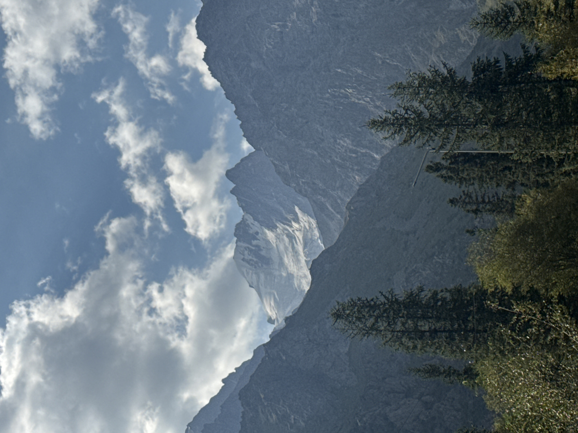 The image depicts a mountainous landscape with a snow-capped peak visible in the distance. The foreground features lush green trees and foliage. In the mid-ground, a tall lamp post stands amidst a cluster of evergreen trees. The sky is partly cloudy, with a mix of bright blue patches and white clouds. The mountains are rugged and appear to be made of dark rock. The overall scene suggests a natural and scenic environment, possibly a rural or wilderness area. The location is identified as Unknown, Kyrgyzstan.