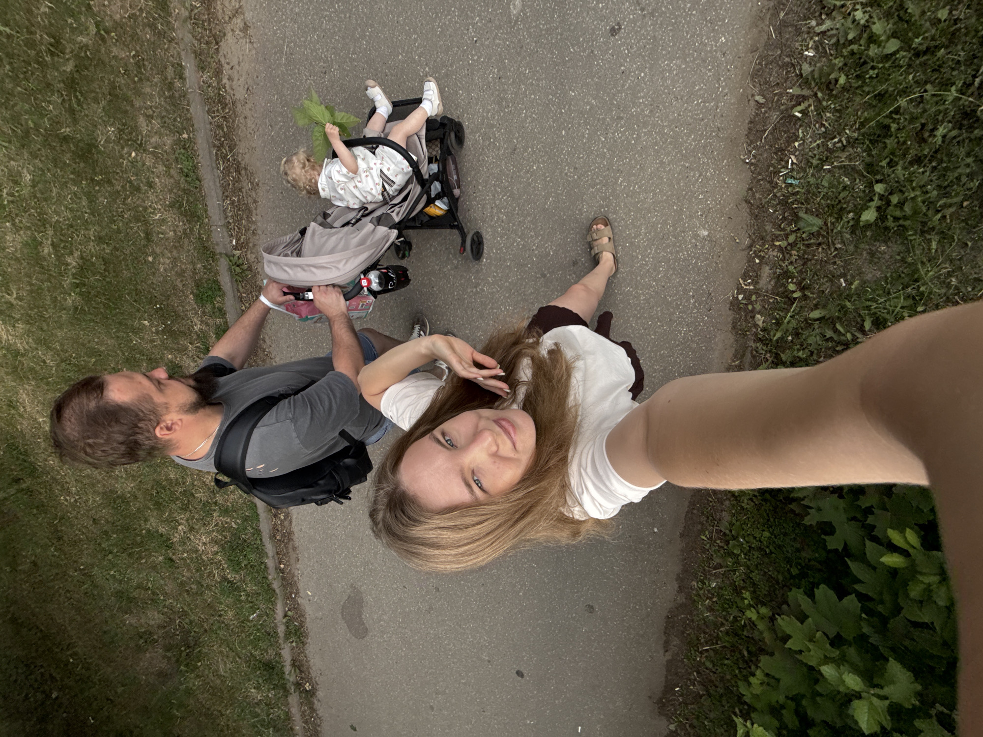 An overhead, slightly tilted shot shows a family on a paved path next to a grassy area. A man with a beard and short hair wears a grey t-shirt and a black backpack. He is pushing a light-colored stroller with a child in it. The child has blonde curly hair and wears a white dress with red floral accents. The child is holding a large green leaf. A woman with long brown hair and a white t-shirt is in the foreground, her arm extended as if taking the photo. She wears sandals. The path is asphalt, and the grassy area has various plants and some scattered debris. The scene appears to be outdoors in a park or a recreational area.