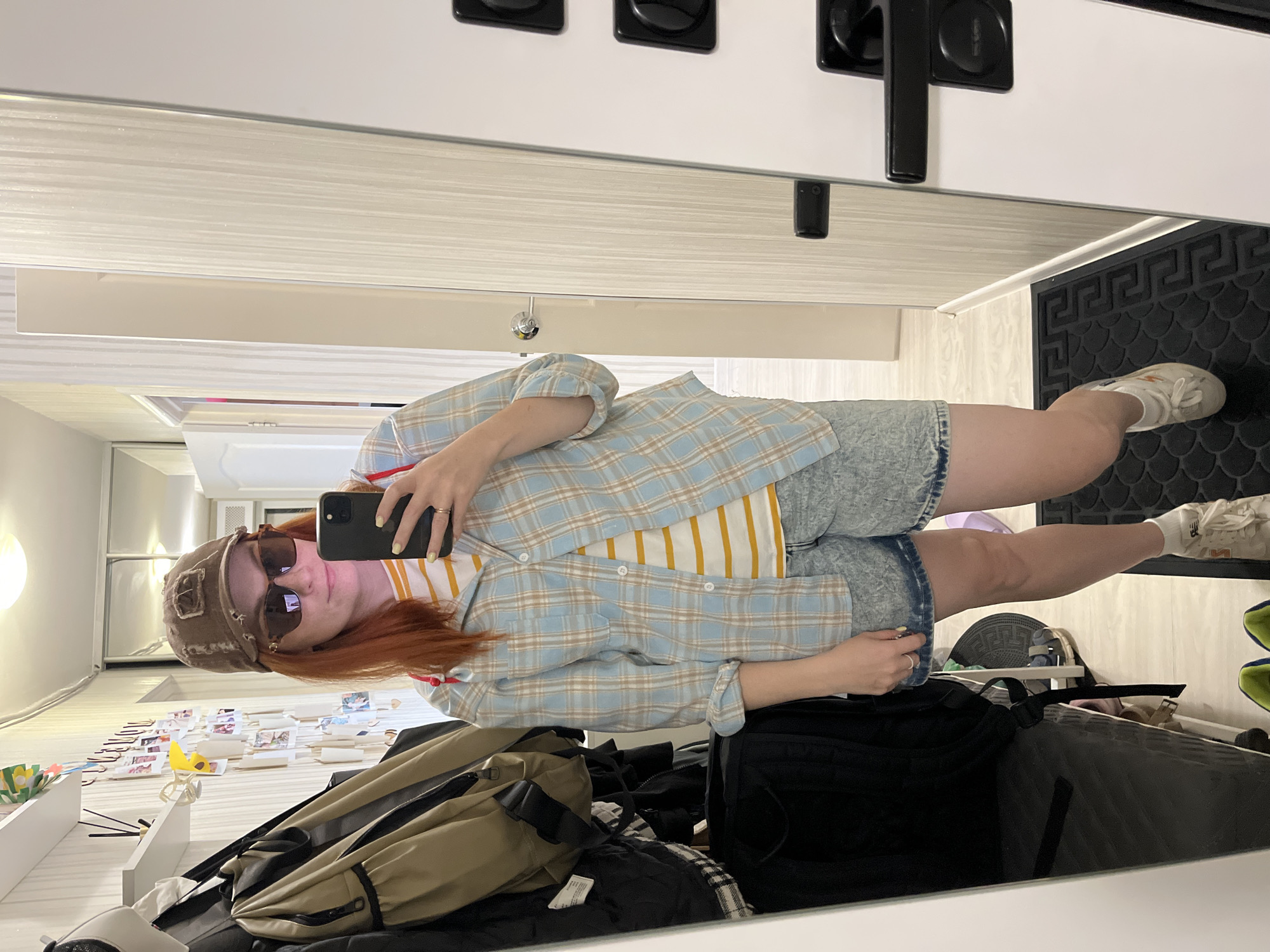The image shows a young Caucasian woman with reddish-brown hair, wearing a brown baseball cap, sunglasses, a blue and tan plaid shirt over a yellow and white striped t-shirt, and acid-wash denim shorts. She is standing in a hallway and taking a selfie in a full-length mirror. The hallway has light-colored, striped wallpaper and a white door with a black handle. To the left of the woman, there is a coat rack with several bags hanging on it. The floor is tiled and there is a black mat with a Greek key pattern in front of the mirror. The scene appears to be in a residential setting, possibly an apartment in Chaikovsky, Russia, given the context provided.