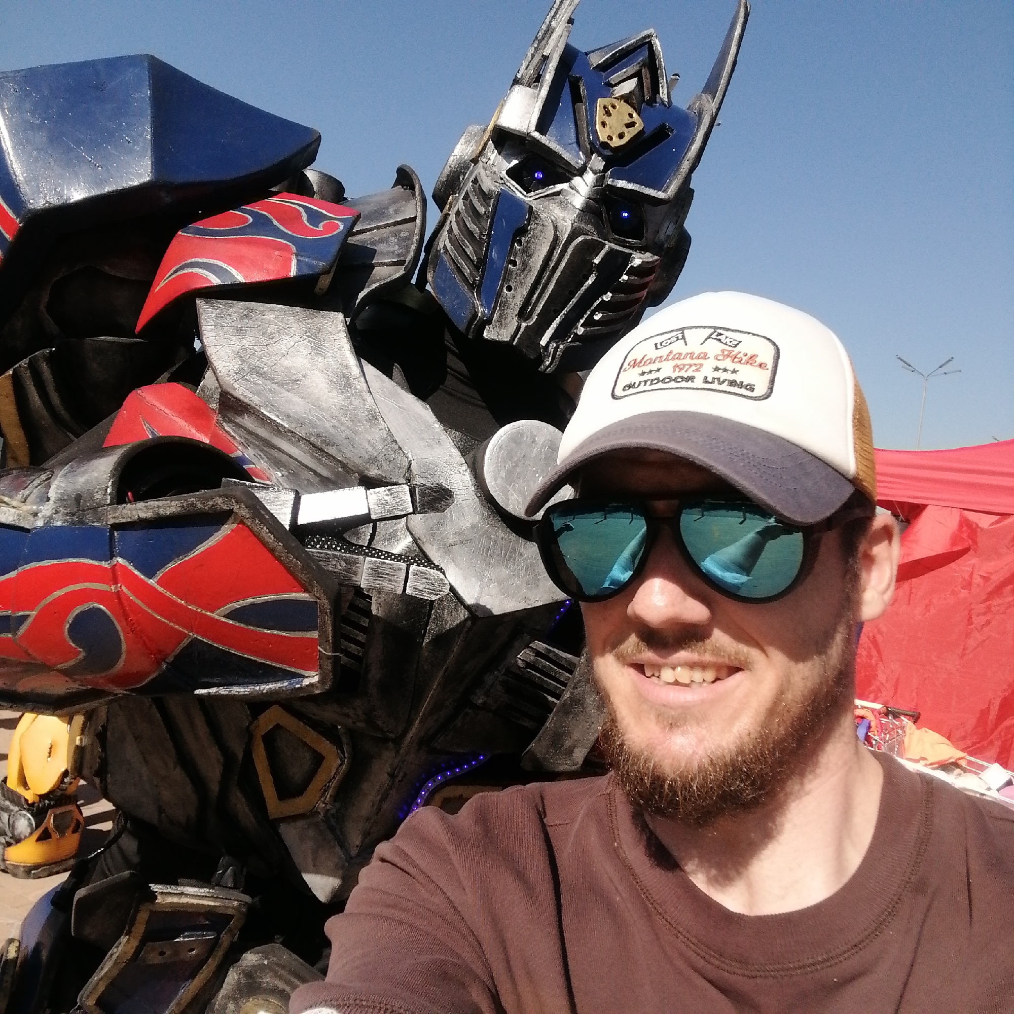 An outdoor daytime scene in Трубачеевка, Russia, features a man in the foreground positioned as if taking a selfie with a robot costume behind him. The man has a beard, fair skin, and is smiling. He wears a white and tan baseball cap with a patch displaying "Montana Hike *** 1972 *** OUTDOOR LIVING," blue mirrored sunglasses, and a brown long-sleeved t-shirt. To his left and behind him stands a robot costume, predominantly silver-grey with blue and red flame-like accents. The robot's head features glowing blue eyes and a gold-colored emblem on its forehead. The background consists of a clear blue sky, a red canopy or tent structure on the far right, and a street light pole visible in the distance.