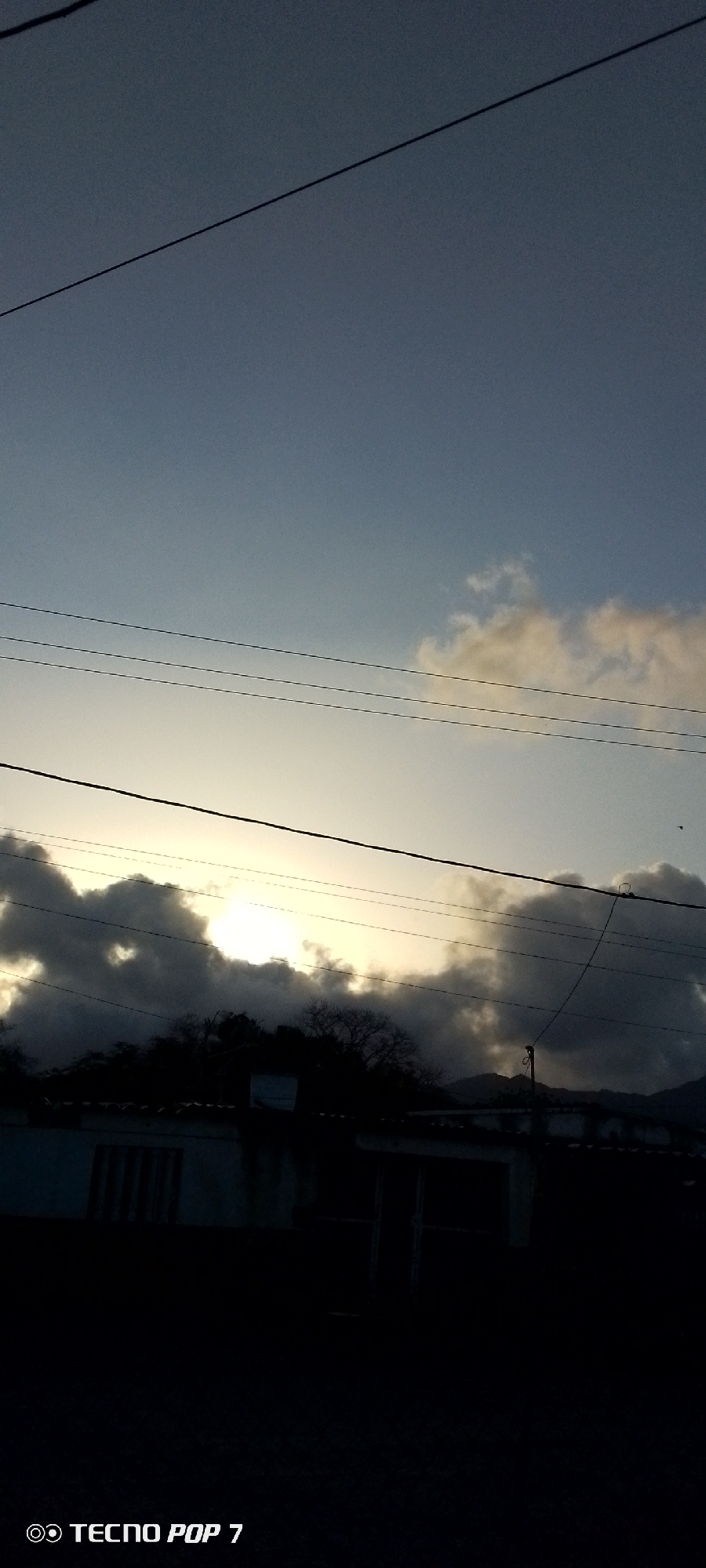The image shows a sky scene taken in Altagracia, Venezuela, at what appears to be dusk or dawn. The sky is light blue fading to a brighter horizon obscured by dark clouds and trees. The sun is trying to break through the clouds. Overhead power lines stretch across the frame. In the foreground, the silhouette of a building can be seen. A mountain range is faintly visible in the background.