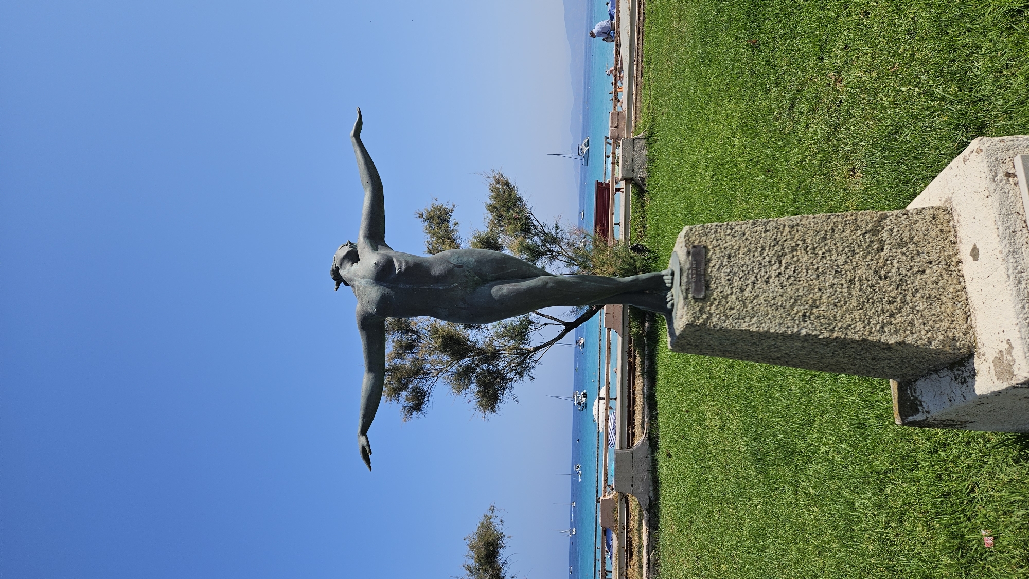 The image shows a bronze statue of a female figure with arms outstretched, standing on a stone pedestal. The statue is positioned in the foreground, with a backdrop of the blue sea, sailboats, and a clear blue sky. A grassy area with trees is visible in the middle ground, and a paved walkway with benches runs along the coastline. The statue appears to be located in a park or public space in L'Île-Rousse, France, as indicated by the inscription "ONDINE" on the pedestal, which is a common reference to nymphs associated with water. The lighting suggests it is a sunny day.
