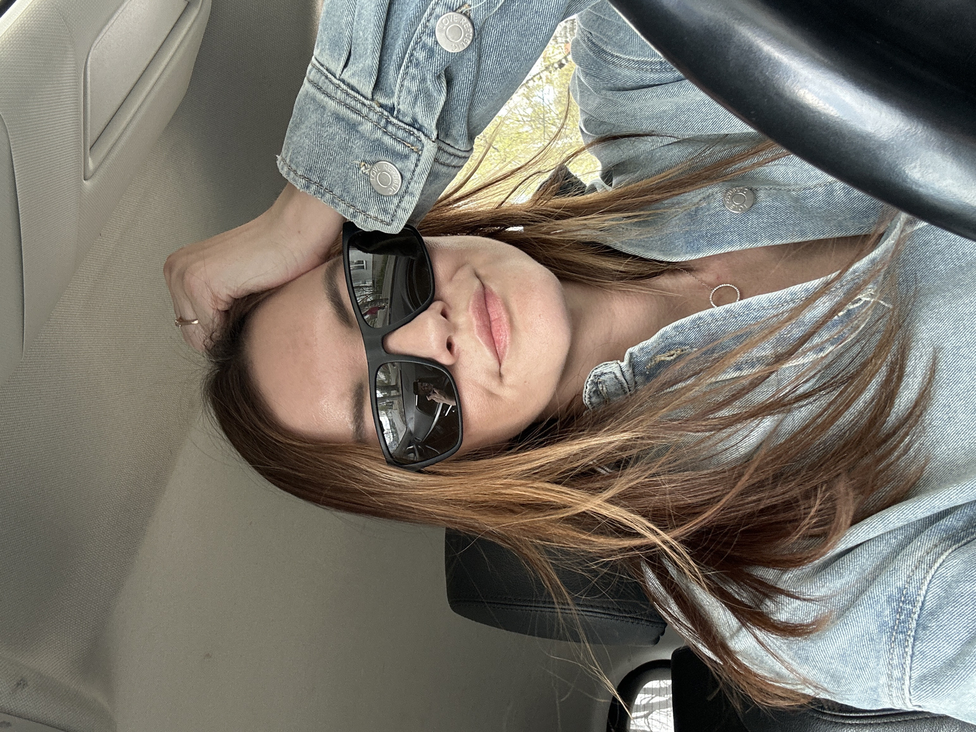A woman with long, brown hair and sunglasses is in a car, wearing a denim jacket and a circular pendant necklace. Her left hand rests on her head. The interior of the car is visible, including the steering wheel, dashboard, and door panel. Reflections can be seen in the sunglasses. The location in Chaikovsky, Russia is not discernible from the image.