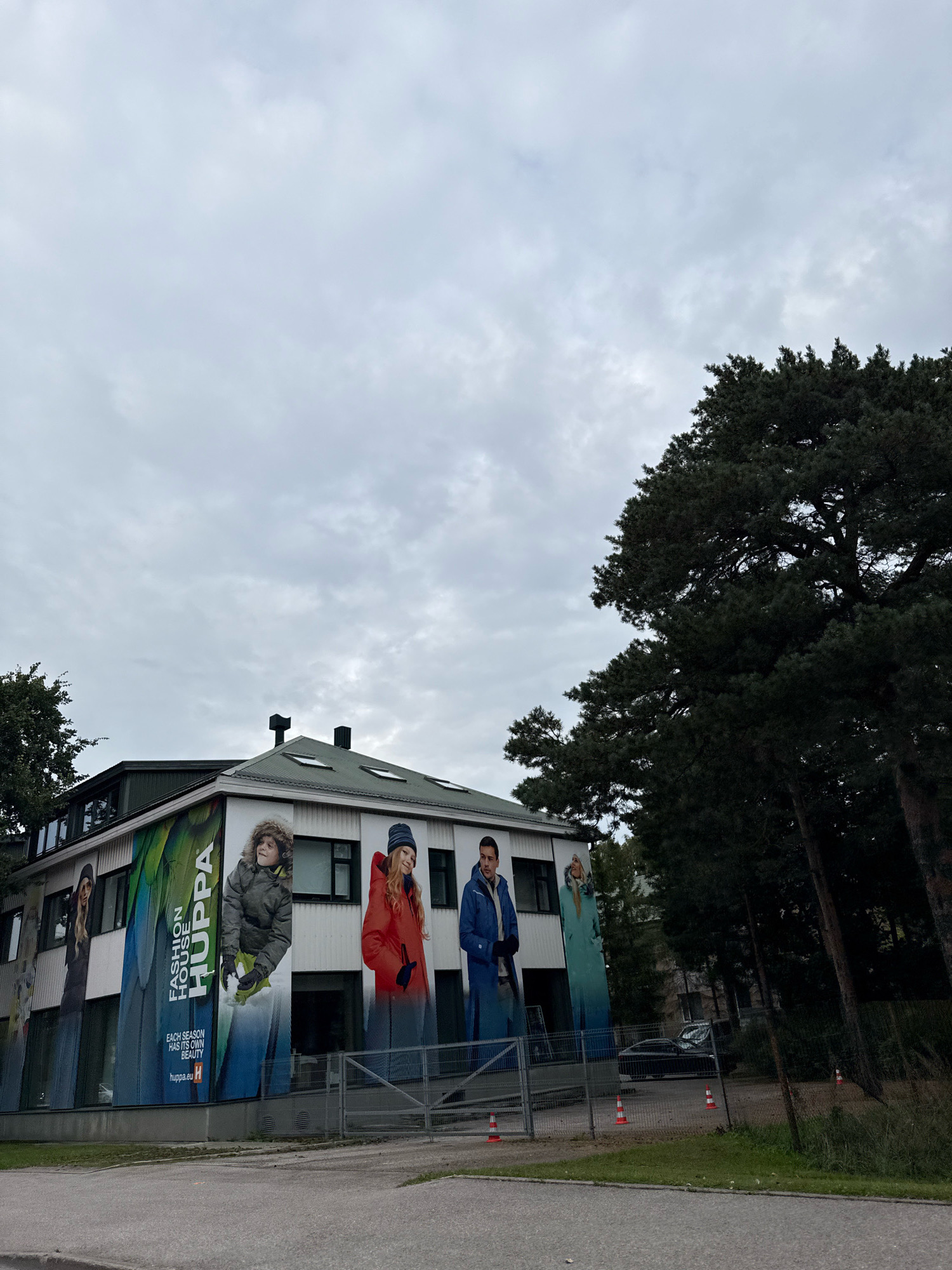 The image shows the "Fashion House Huppa" building located on Lootsi Street, Tallinn, Estonia. The building is a two-story structure with a light-colored facade adorned with large banners featuring models wearing winter clothing. The banners display the brand name "HUPPA" and the tagline "EACH SEASON HAS ITS OWN BEAUTY". The building is surrounded by trees, including tall pine trees on the right side of the image. A grey fence runs along the front of the property, and several orange and white traffic cones are visible on the asphalt in front of the fence. A dark-colored car is parked behind the fence. The sky is overcast.