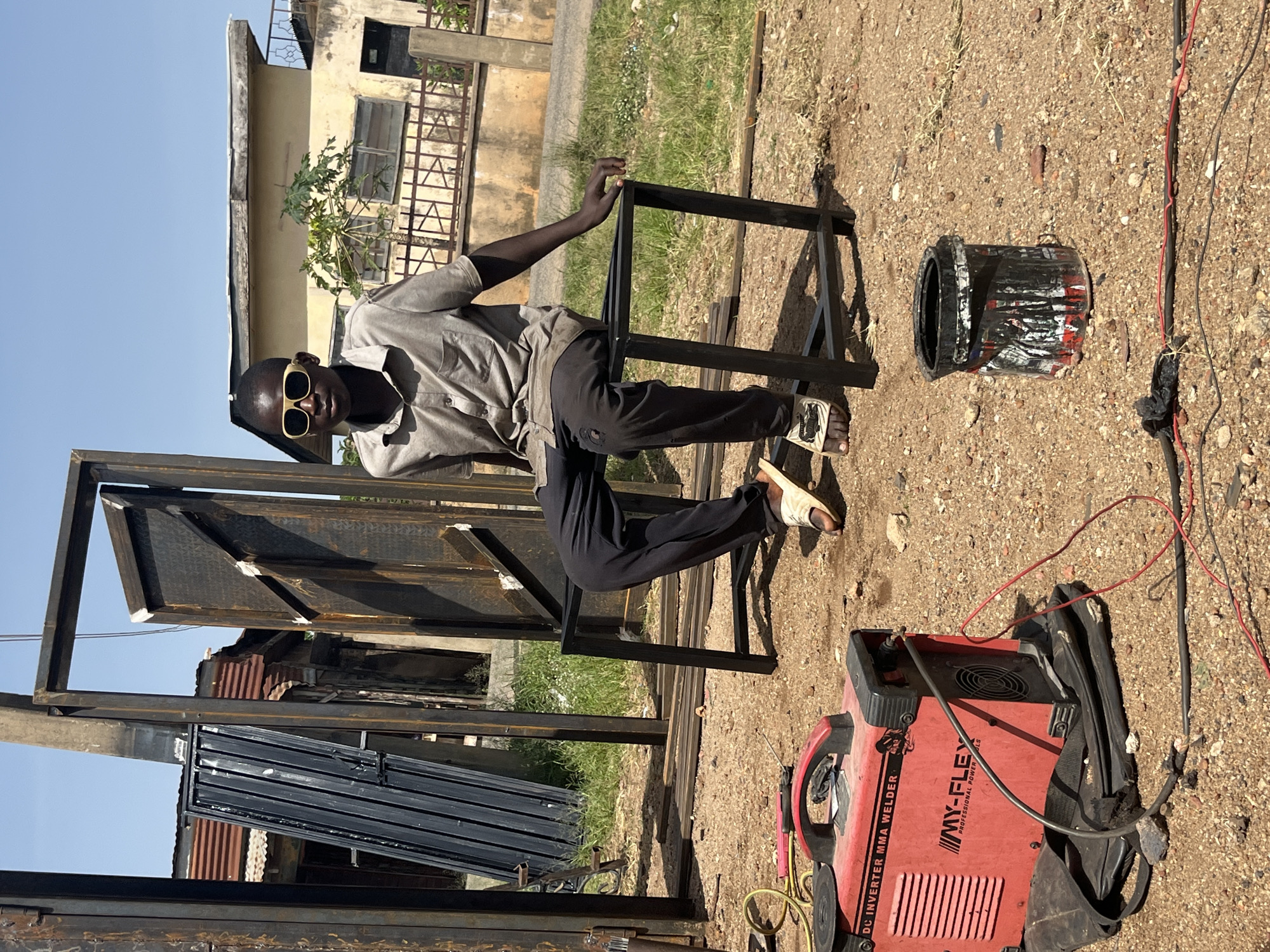 A young male wearing sunglasses, a gray collared shirt, and dark pants sits on a black metal bench in a dusty outdoor area. He is surrounded by metal framing for doors or gates, with a black ribbed metal gate leaning against a frame to his left. To his right, a weathered yellow building with a corrugated metal roof stands in the background, behind a metal fence. In the foreground, a red DC inverter MMA welder and a black paint can are visible on the ground. The scene appears to be in a workshop or construction site in Jos, Nigeria, given the presence of welding equipment and the architectural style of the building. The lighting suggests it is a sunny day.