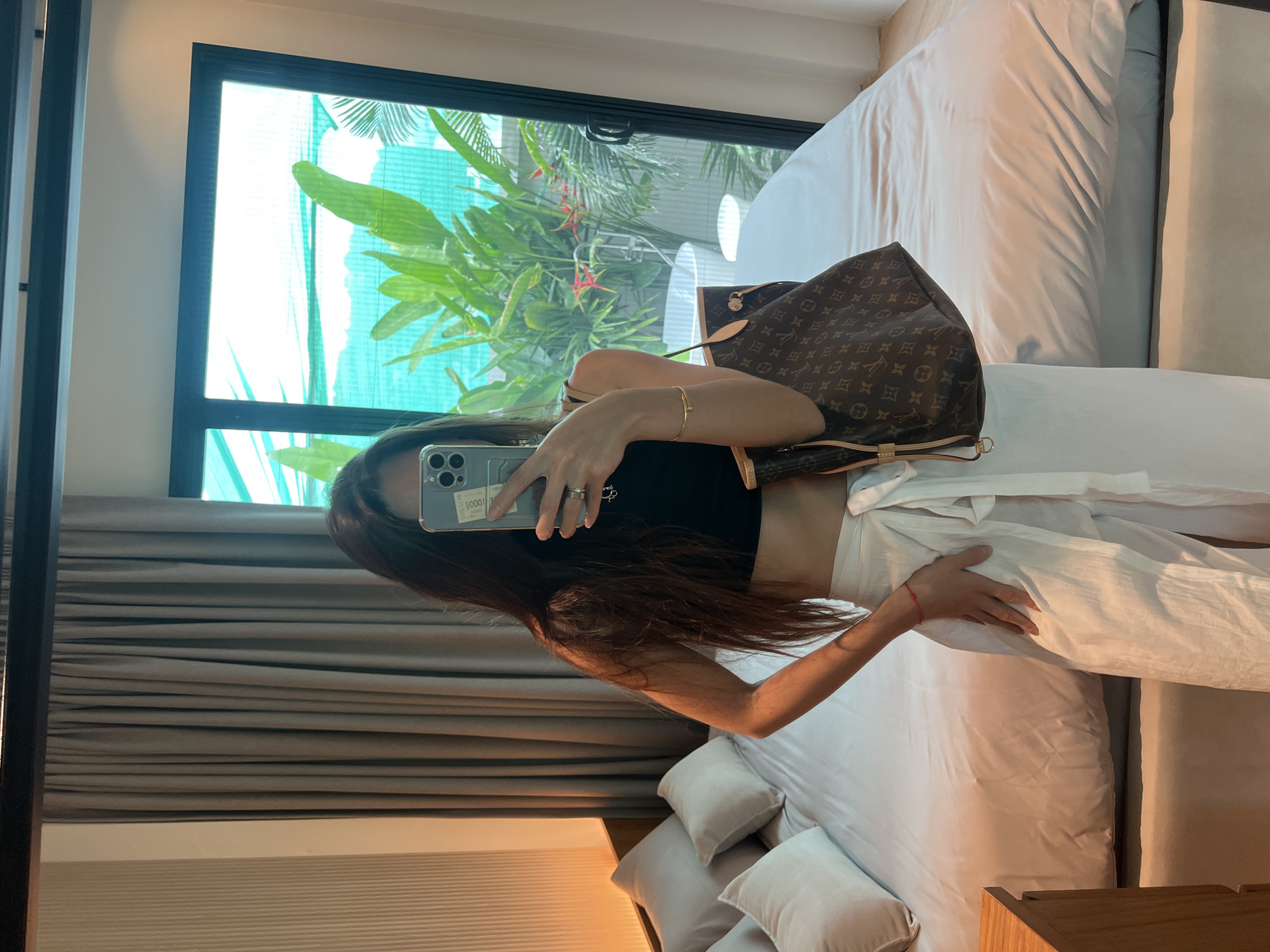 A person is shown from the thighs up, holding a cell phone up to take a mirror selfie while indoors. The person has long brown hair and wears a black crop top, white pants, and a gold bracelet on the right wrist. A Louis Vuitton purse hangs over the person’s left shoulder. The room includes a large window with a view of tropical trees, a gray curtain covering the left side of the window, and a large bed with a white comforter and multiple pillows. The location is in the city of Unknown, Unknown.