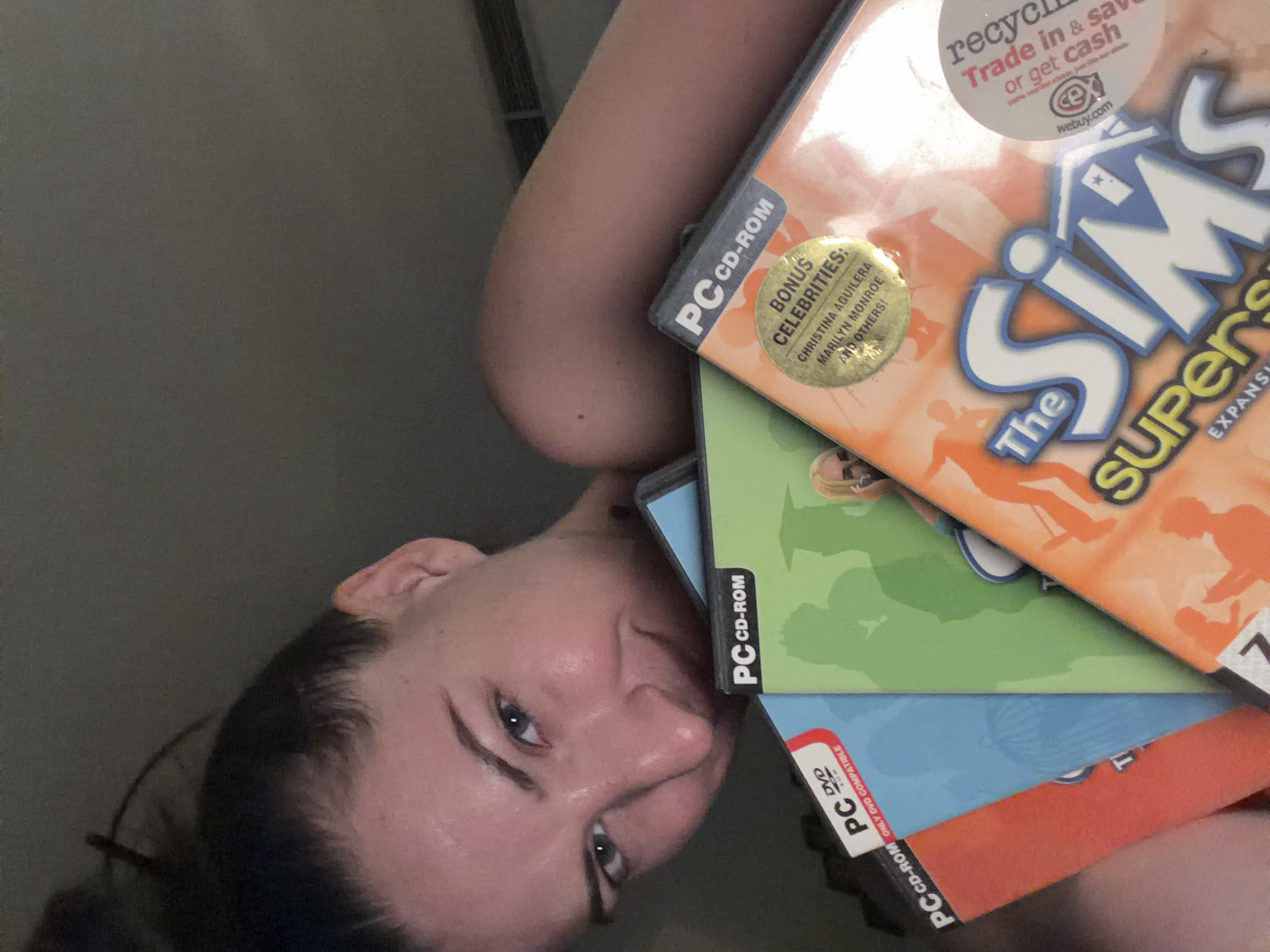 A woman is holding several PC game cases in front of her face, obscuring the lower half of her face. She is looking directly at the camera with a slight smile. The game cases are stacked and tilted, with the titles "The Sims Superstars" and "PC CD-ROM" visible. The background is dark and out of focus, suggesting an indoor setting. There is no information to suggest the image is located in Sharm El Sheikh, Egypt.
