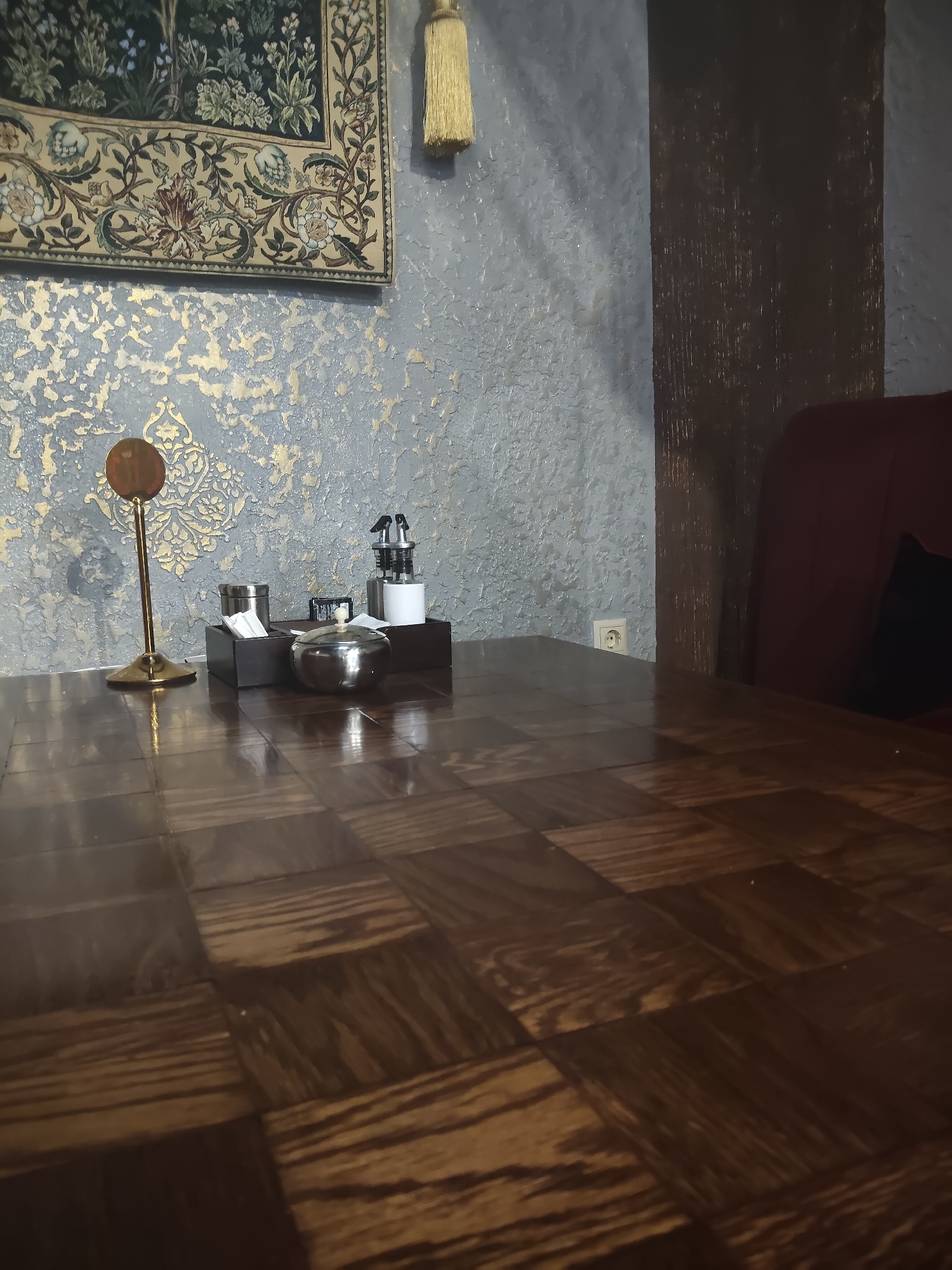 The image depicts an indoor scene, likely a restaurant or cafe, with a strong emphasis on the textured wallpaper and the polished wooden tabletop. The wallpaper is a mottled grey with an overlay of gold metallic detailing, creating a distressed and ornate effect. A woven tapestry with a floral motif and a decorative gold tassel hangs on the wall in the upper left quadrant.

On the tabletop, which has a parquet pattern of dark wood, are several items. To the left, a thin, gold-colored stand holds a circular, bronze-colored disc. To the right of this stand is a dark wooden tray containing a metallic sugar bowl with a lid, a stack of white napkins, and what appear to be sugar packets or similar small items. Behind these, in a white dispenser with two spouts, are likely condiments such as oil and vinegar. A deep red upholstered chair is visible on the right side of the frame, with a black pillow resting on it. An electrical outlet is visible on the wall to the left of the chair.

The image is a still life, with no people or discernible actions taking place. The lighting suggests an interior environment, possibly with artificial lighting contributing to the sheen on the wooden table. The overall impression is one of a slightly formal or elegantly decorated dining space. The provided location of Chekhov, Russia, is an external detail and not visually verifiable from the image itself.