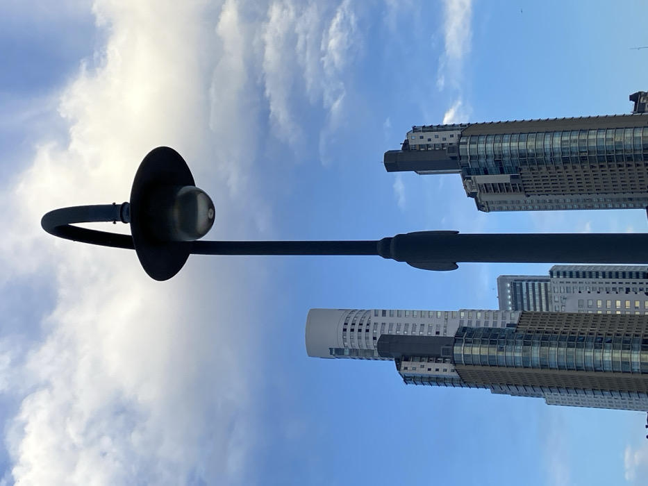 The image captures an upward-looking perspective of an urban environment during the daytime. A prominent street light pole, dark in color, extends diagonally from the lower left to the upper right of the frame. Its distinct curved arm holds a dark, disc-shaped fixture with a clear or frosted bulb visible at its center, positioned in the upper left quadrant.
Behind and to the right of the street light, several tall, modern buildings are visible against a bright blue sky. These skyscrapers feature multiple stories with numerous windows, some displaying reflections. One building in the mid-right has a distinctive curved facade with horizontal window bands. Another building, further to the right, appears to be taller with a more rectangular, blocky top section. The structures suggest a developed city landscape.
The sky is a vibrant blue, partially covered by scattered, soft white clouds, indicating clear weather. The lighting suggests it is daytime, likely mid-day or late afternoon. There is no visible human activity or text in the image. Given the provided context, the scene is located in Buenos Aires, Argentina. The overall impression is one of modern architecture contrasting with the natural sky, framed by an urban utility fixture.