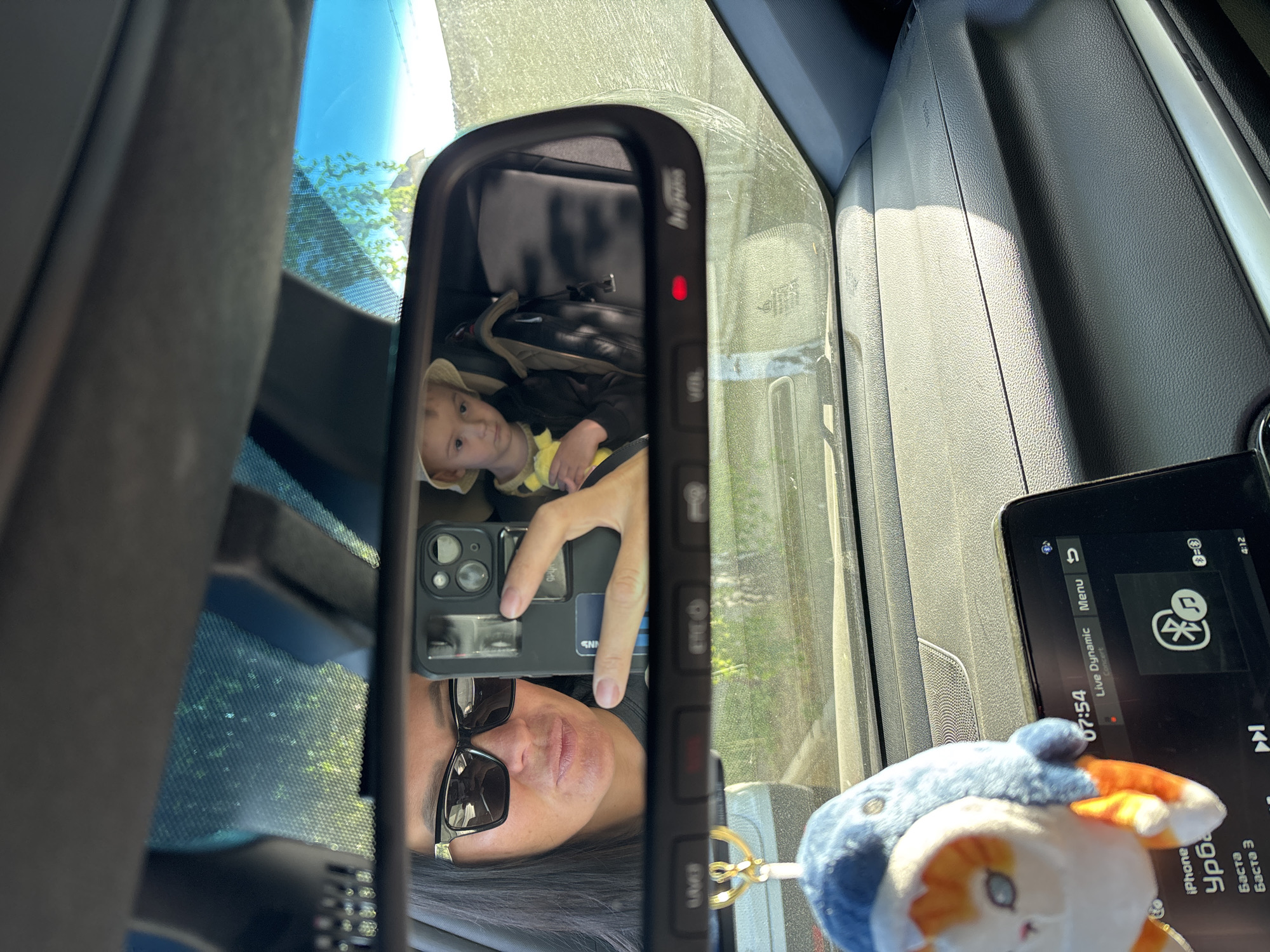 The image appears to be taken from inside a vehicle, likely a car. The rear-view mirror reflects a child in a car seat, along with the person holding a smartphone taking the photo. The phone has a credit card attached. The dashboard is visible with a display screen showing time, audio information (iPhone Урба, Баста, Баста 3), and audio settings. A blue and orange stuffed animal dangles near the screen. Outside, there appears to be greenery. The image was probably taken somewhere in Chaikovsky, Russia.