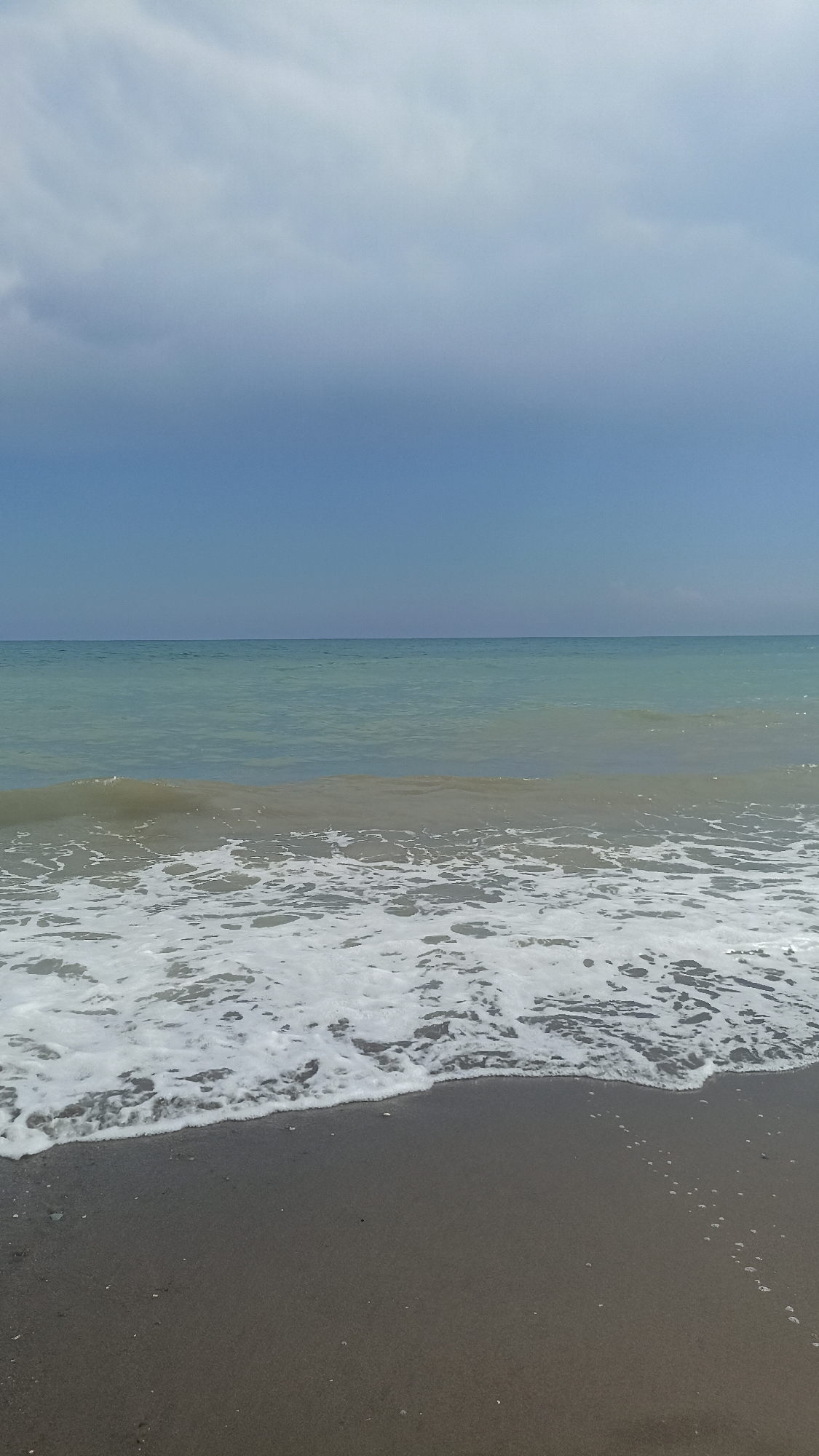 The image depicts a serene beach scene with a calm sea under a partly cloudy sky. The foreground shows dark sand wet from the receding tide, with a line of white foam marking the edge of the water. Gentle waves are breaking on the shore, creating a frothy texture. The sea itself is a tranquil turquoise, stretching to the horizon where it meets a pale blue sky. A band of soft, diffused clouds occupies the upper portion of the sky, obscuring direct sunlight and lending a muted quality to the scene. The overall impression is one of peacefulness and natural beauty. This scene is consistent with many beaches found along the coastline of Mezitli, Turkey, known for its Mediterranean shores and clear waters.
