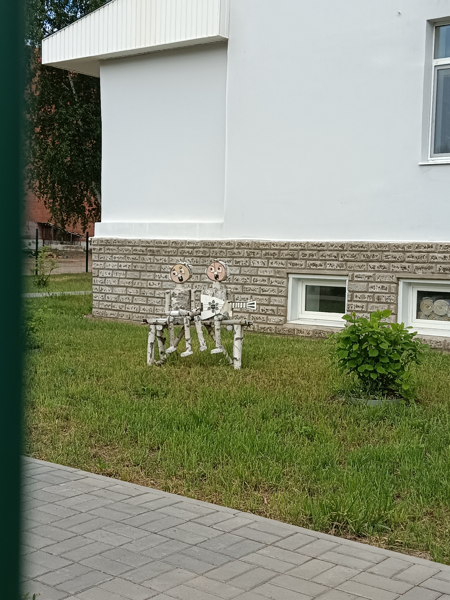 This outdoor daytime image captures a whimsical display in front of a light-colored building, likely in Noviy, Russia. Two decorative figures, crafted from natural wood branches and logs, are seated on a small, rustic bench on a neatly trimmed grassy lawn. The figures have round, flat wooden faces with painted features, including large, open mouths and wide eyes, giving them surprised or cheerful expressions. The figure on the right appears to hold a small, patterned object.
Behind the figures, the building features a lower section with a grey, textured facade resembling brick or stone, and two small, white-framed rectangular windows. Above this, the wall is smooth and white, topped by a white overhang with vertical paneling. To the right of the figures, a small, vibrant green bush grows in the grass.
The foreground shows a grey paved walkway with a herringbone pattern, running along the edge of the lawn. A dark green fence or barrier is partially visible on the far left of the frame. In the background, beyond the fence and building, trees with green foliage and a hint of another building are visible. The lighting is bright and even, suggesting a clear day. No discernible text is visible in the image.