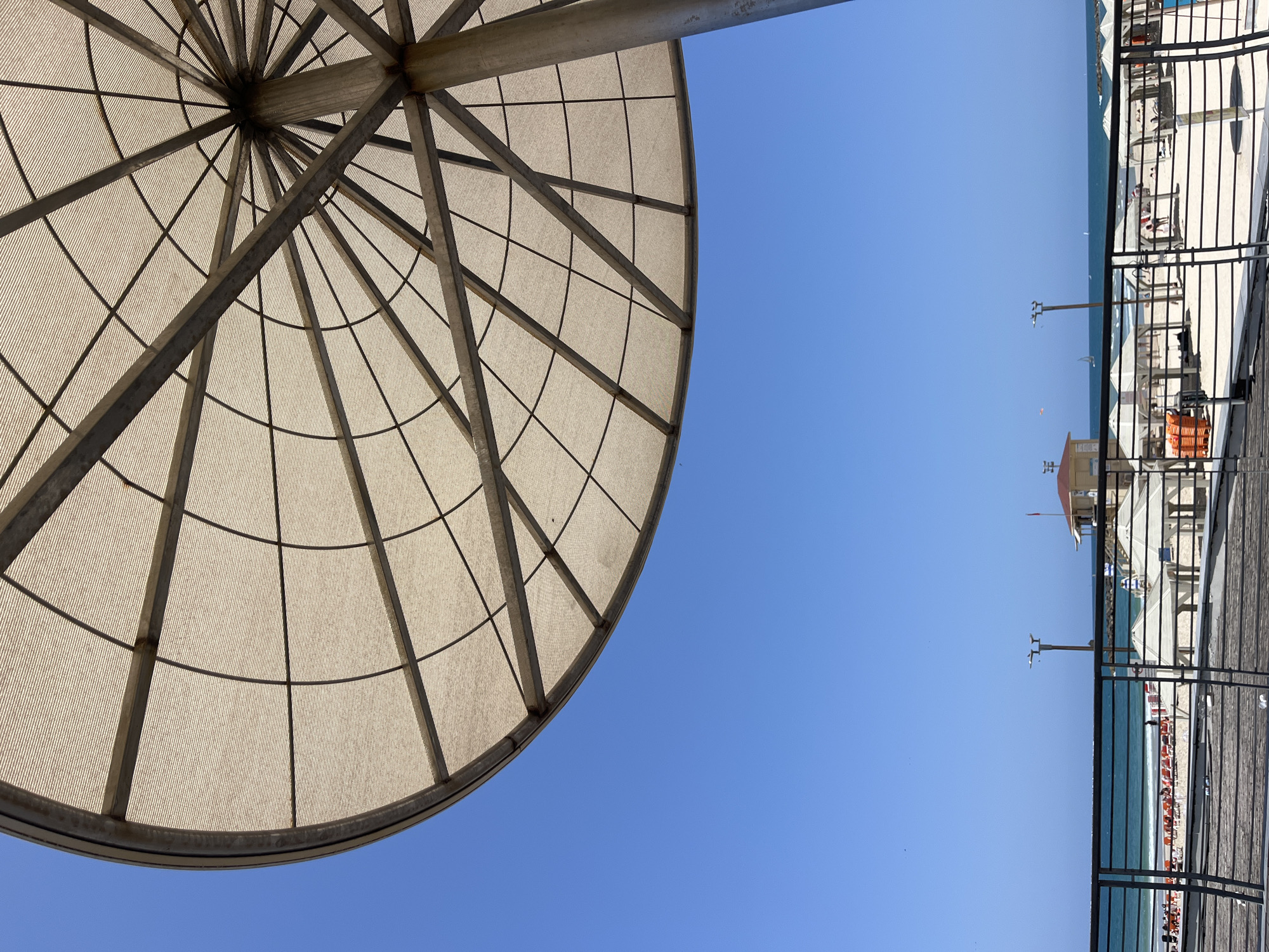 The image, correctly oriented, depicts a vibrant beach scene under a clear blue sky, viewed from an elevated perspective, likely a boardwalk or promenade.
In the upper left foreground, a significant portion of a large, circular shade structure is visible. It features a robust metallic grid framework supporting a light-colored, translucent fabric or mesh covering, which casts a patterned shadow. A thick metal support beam extends from the top right of this structure.
The expansive blue sky fills the upper right and central parts of the frame, indicating a sunny day with no clouds. Below the sky, the serene turquoise waters of the sea stretch towards the horizon, meeting a wide, sandy beach. The beach is well-populated with numerous beach chairs or loungers, some appearing orange, and several beach umbrellas, suggesting active use by sunbathers or beachgoers, whose small figures are faintly visible. A distinctive lifeguard stand, featuring a red roof and white base, is situated on the sand.
A black metal fence or railing runs along the right side of the image, separating the viewing platform, which includes a dark wooden boardwalk, from the beach below. The bright, even lighting points to mid-day or early afternoon. No discernible text is visible in the image. The scene conveys a relaxed, sunny atmosphere typical of a summer day on a coastal resort, consistent with the location context of Tel-Aviv, Israel.