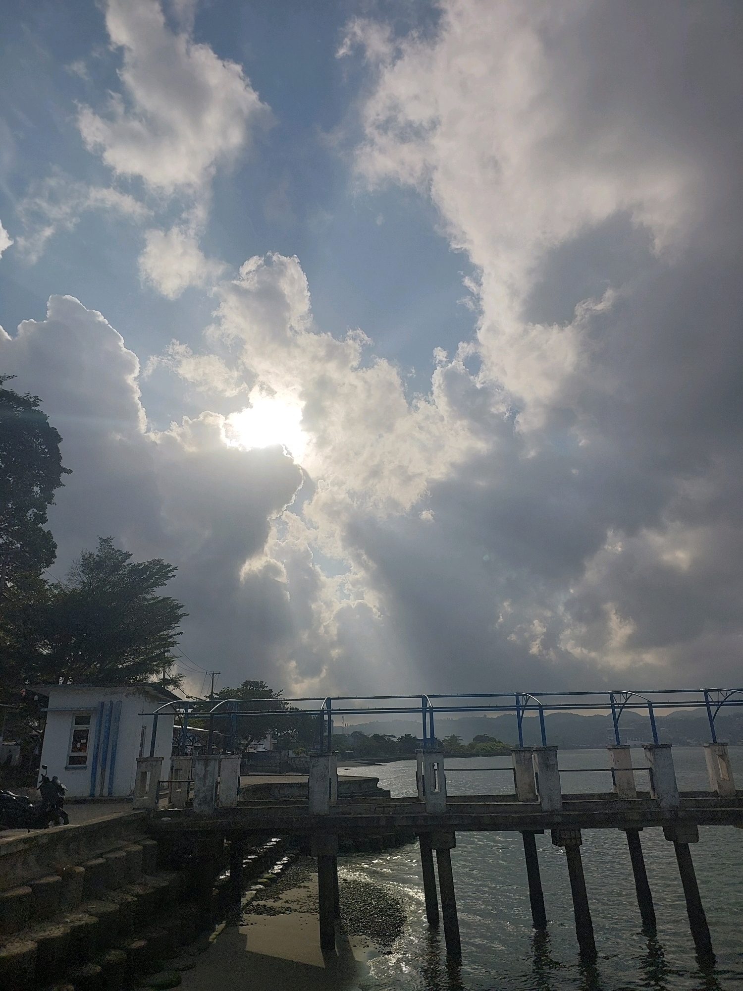 The image depicts a waterfront scene in Ambon, Indonesia, with a pier extending over the water. The sky is dramatic, with the sun breaking through a dense cloud cover, casting rays of light downwards. The pier is constructed of concrete and has blue railings. To the left of the pier, a small white building with blue trim is visible, along with a parked motorcycle. The water is choppy and reflects the light from the sky. In the background, hills and trees can be seen on the horizon. The overall impression is one of a dynamic and somewhat moody natural environment at the edge of an urban area.