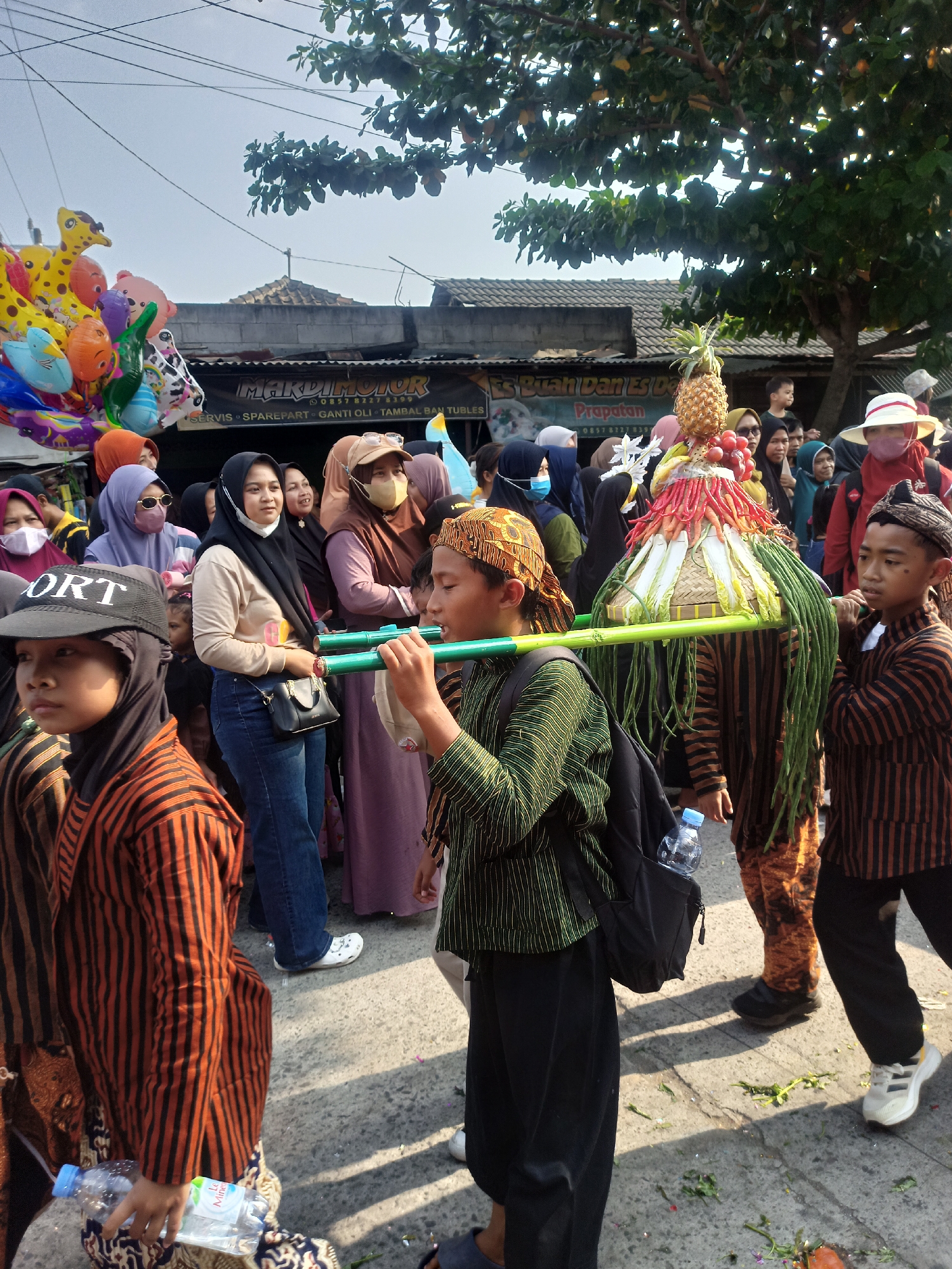 This is a medium shot of a street procession in Saren, Indonesia. The procession appears to be a cultural or religious event. Several people, including children and adults, are visible, many wearing traditional clothing. A prominent float, decorated with fruits and vegetables, is being carried by two young boys. The background shows buildings and trees, suggesting an outdoor, urban setting. The overall atmosphere appears festive, with a crowd of onlookers and a bright, sunny day.