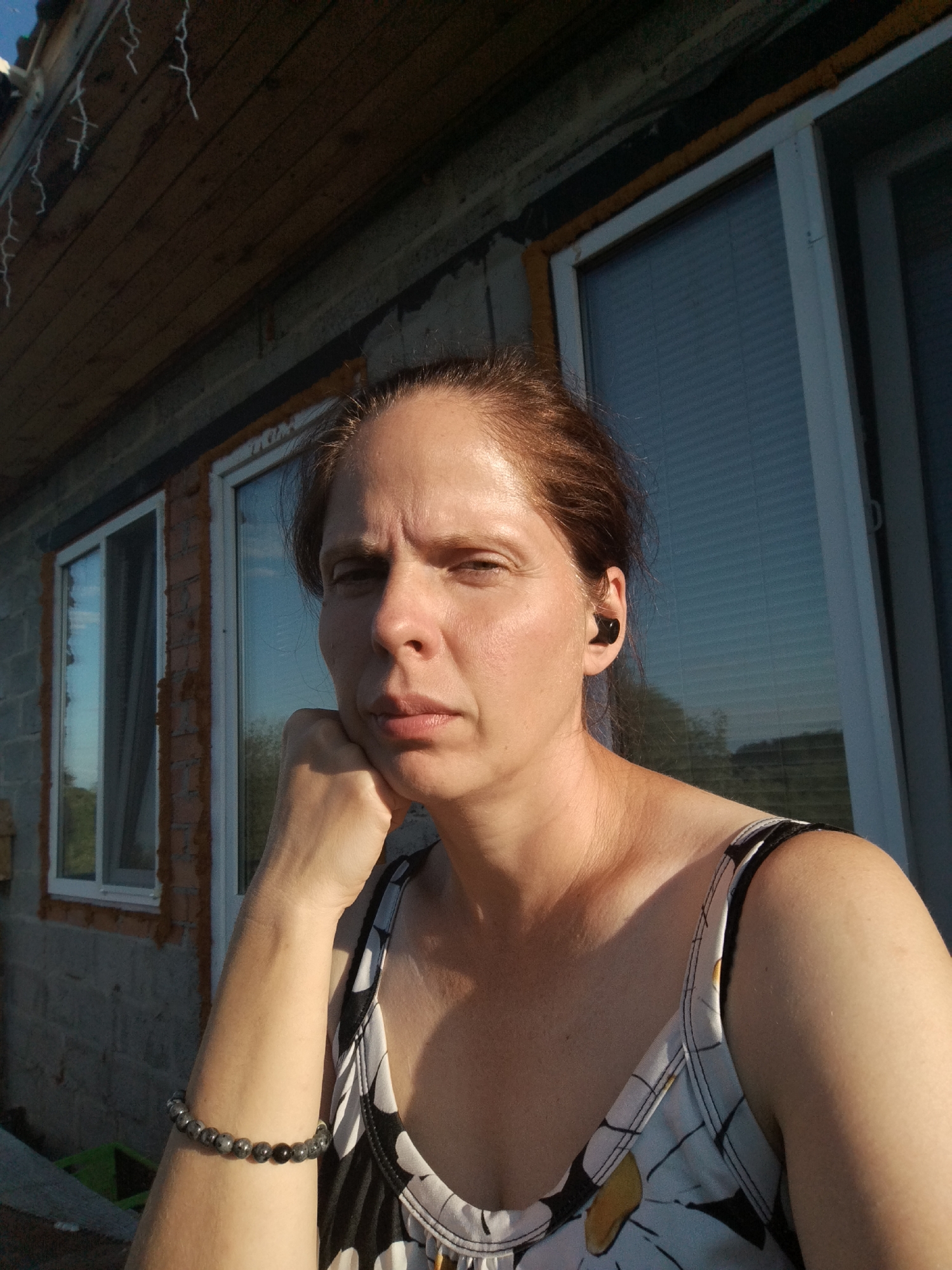 The image shows a woman with brown hair and fair skin, wearing a black and white floral print tank top and a beaded bracelet on her left wrist. She has a black earbud in her right ear and is resting her chin on her left hand. She is positioned in front of a building with unfinished brickwork and windows. The scene appears to be outdoors during the day, with sunlight casting shadows. The location is identified as Zavodoukovsk, Russia.
