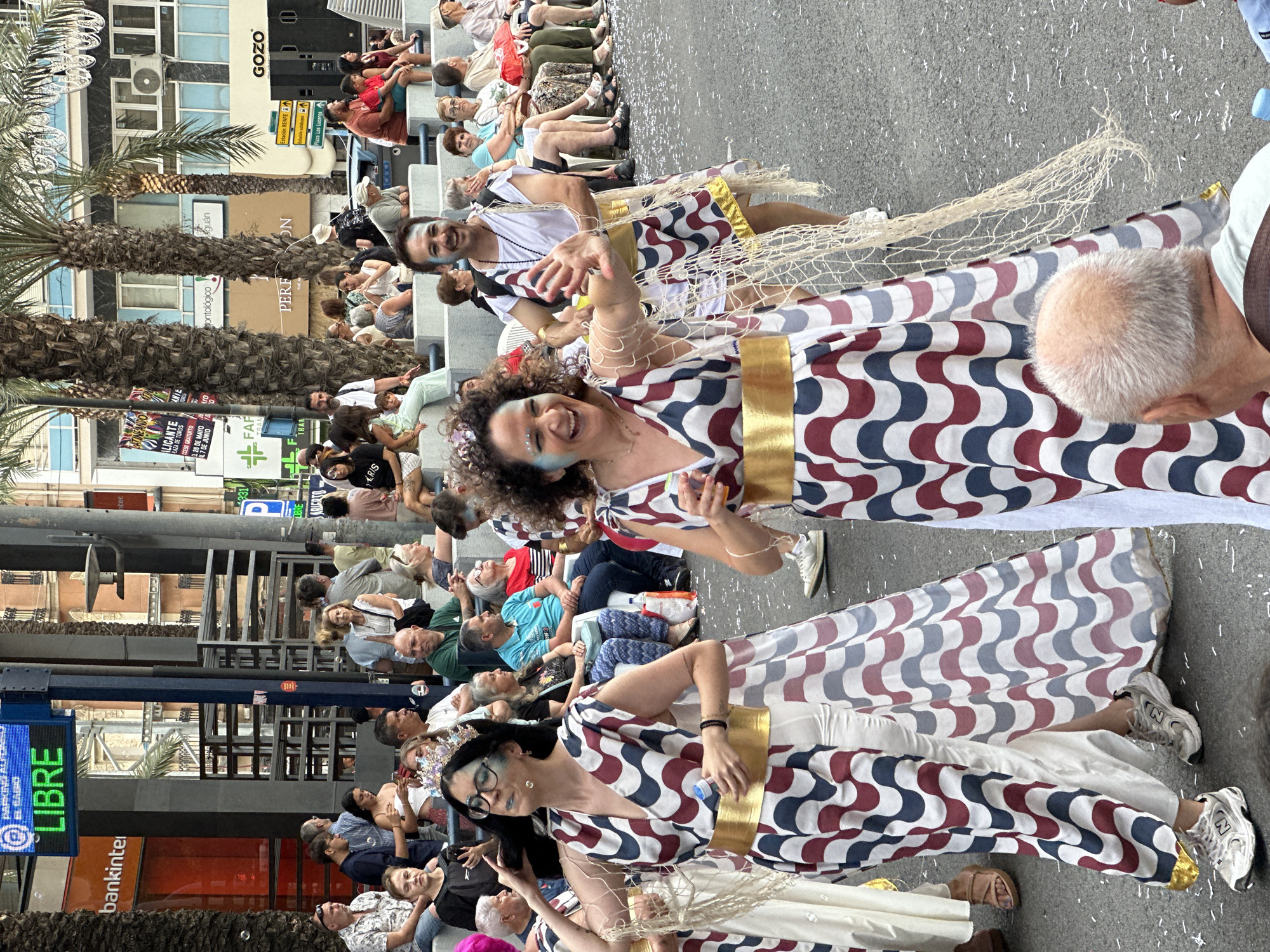 The image depicts a daytime outdoor street scene in Alicante, Spain, likely during a festive event or parade. In the foreground, three individuals in distinct costumes are actively participating. The central figure is a woman with curly hair, a broad smile, and blue facial makeup, wearing a long dress with a repeating wavy pattern in dark red, dark blue, and light grey on a white background. She has a wide gold belt and a light-colored net-like fabric draped over her shoulders. Her left arm is raised as if waving. To her left, another woman in an identical patterned dress, gold belt, and blue facial makeup is visible, also smiling slightly while holding a small blue bottle. She wears glasses and a crown-like headpiece. Both women wear athletic shoes. Partially visible to the right and behind the central woman is a man in a white tunic with a similar patterned fabric draped over it, a gold belt, and blue facial makeup, also smiling widely. The ground beneath them is dark asphalt scattered with numerous small white paper or confetti pieces.
In the midground and background, numerous spectators are seated on tiered bleachers or elevated platforms on both sides of the street, observing the event. They are dressed in casual attire. Behind the spectators on the left, a "Bankinter" building is visible, along with a blue digital sign displaying "PARKING ALICANTE EL BARRIO" and "LIBRE" in green text. Several tall palm trees line the street. On the right side of the street, various buildings are visible, including one with "GOZO" written on it and a street sign indicating "Estación Renfe" and "Plaza Luceros." Overhead, decorative string lights are strung across the street. The overall lighting is bright but diffused, suggesting an overcast day or shaded area.