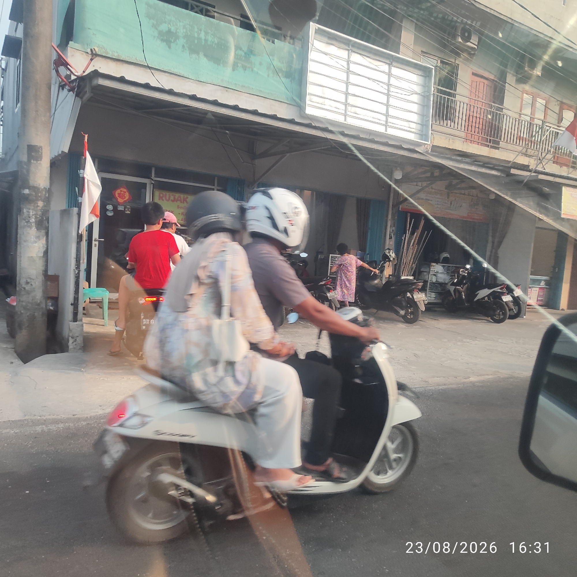 The image depicts a street scene in Tanjung Pandan, Indonesia, on August 23, 2026, at 16:31. Two individuals are riding a white scooter. The person in the front is wearing a grey shirt and a white helmet with blue accents. The person behind them is wearing a light-colored patterned top and white pants, with a white handbag on their lap. They are both wearing helmets.
In the background, a building with a green facade and a metal awning is visible. A red and white Indonesian flag is attached to the building. Various businesses appear to be located within the building, with signage that includes the word "RUJAK." Several other motorbikes are parked in front of the establishments. The street surface is asphalt. The overall lighting suggests it is late afternoon.