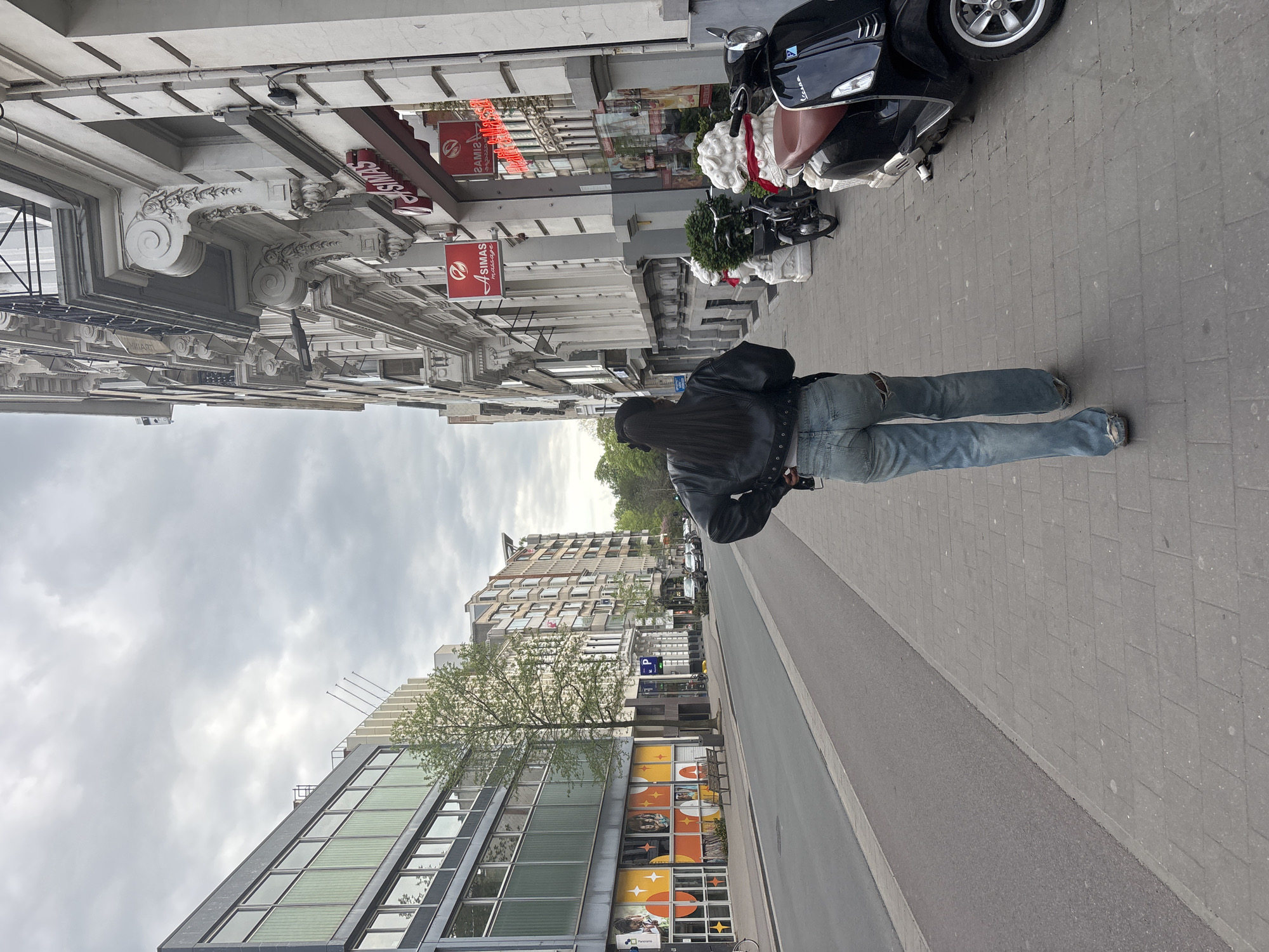 A photograph shows a person walking on a sidewalk in Antwerp, Belgium. The individual is wearing a black leather jacket and blue jeans. A black Vespa-style scooter is parked on the sidewalk nearby. Buildings line the street, with "SIMAS massage" signs visible on one. The sky is overcast. The photo is taken from a low angle.