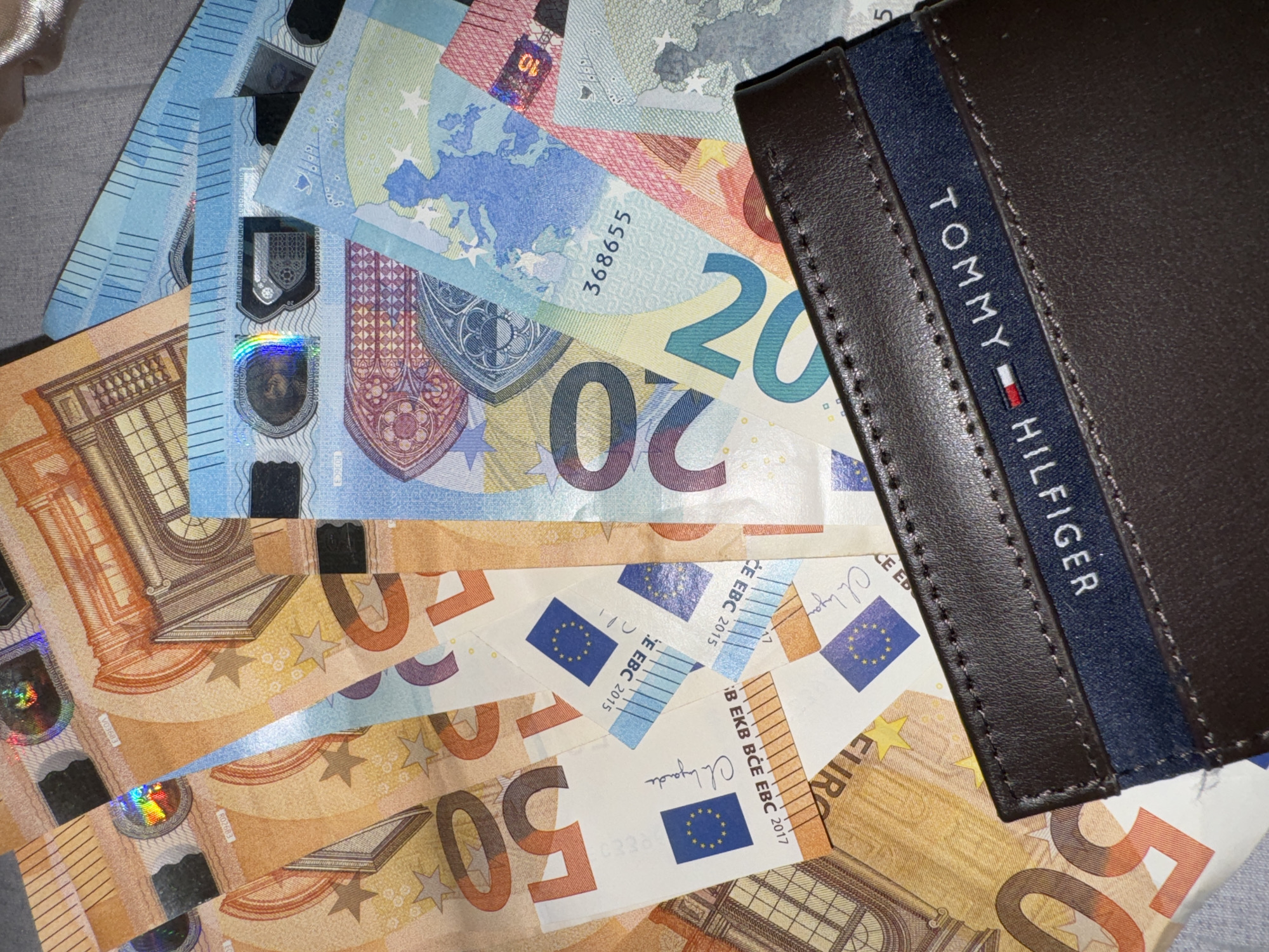 The image shows a close-up of a disorganized pile of Euro banknotes in denominations of 10, 20 and 50 euros. A dark brown Tommy Hilfiger wallet is partially visible on the right side of the image, overlapping the banknotes. The scene is indoors, illuminated by dim light.
