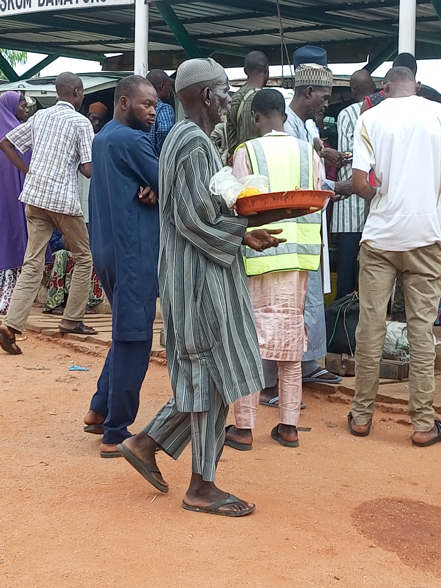 The image shows a group of men standing on a dirt surface, possibly at a market or gathering in Gombe, Nigeria. In the foreground, an elderly man wearing a gray and white striped robe and a gray skullcap is holding an orange tray with some food items. He is looking to his right. To his left, a man in a dark blue robe stands with his arms crossed. Behind them, several other men are visible, some wearing casual attire and others in traditional clothing. One man in the center wears a yellow safety vest over a pink garment. The background shows a covered structure with a white ceiling, and what appears to be a white vehicle. The ground is covered in reddish-brown dirt, with some debris scattered around. The overall scene suggests a public gathering where food may be exchanged or consumed.