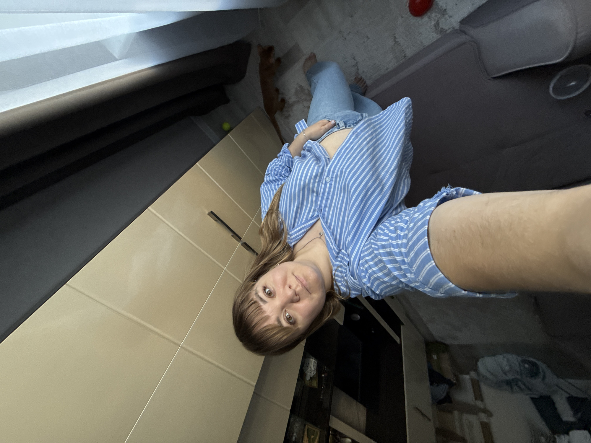 This is a vertically oriented, first-person perspective photograph taken inside an apartment in Stary Oskol, Russia. The subject is a young woman with shoulder-length brown hair and bangs, wearing a blue and white striped button-up shirt and light blue jeans. She is holding the camera with her right hand, extending it to take a selfie. Her expression is neutral.
In the background, a ginger cat stands on a rug, facing away from the camera. To the left, a portion of a beige cabinet is visible. The floor appears to be carpeted in some areas and tiled in others. A yellow ball is on the floor near the cat. A red object, possibly a toy, is also visible on the floor in the upper right portion of the image. The lighting suggests it could be daytime, but the indoor setting makes it difficult to determine the exact time of day or weather. The overall impression is a casual, domestic scene. No visible text is present in the image.