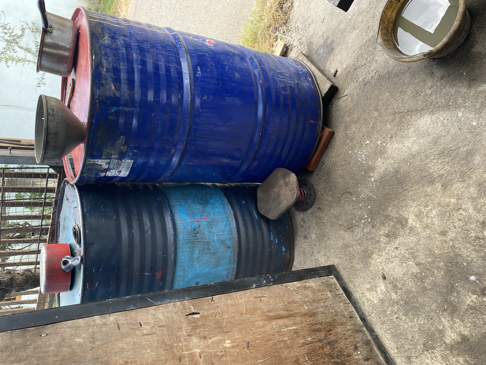 The image depicts an outdoor scene with two large industrial drums and various objects arranged on a concrete surface. A wooden panel, possibly part of a door or barrier, is positioned on the left side of the frame. The two drums are cylindrical and appear to be made of metal. The drum on the left is predominantly black with a blue section, and it has a red lid with a black handle mechanism. On top of this lid, a dark object, possibly a cap or tool, is present. The drum on the right is bright blue and has a red top. Placed on the red top of the blue drum are a dark funnel and a metallic pot with a handle. A small wooden stool, supported by a spring mechanism, is situated between the two drums. In the foreground, a bucket filled with murky liquid is visible on the concrete. The background shows a fence, vegetation, and a building facade, suggesting an industrial or workshop setting. The ground is a rough concrete surface with some debris and patches of dried grass and dirt visible. The overall lighting suggests daylight. The scene does not contain any individuals. The location is identified as the city of Bayu, Indonesia.