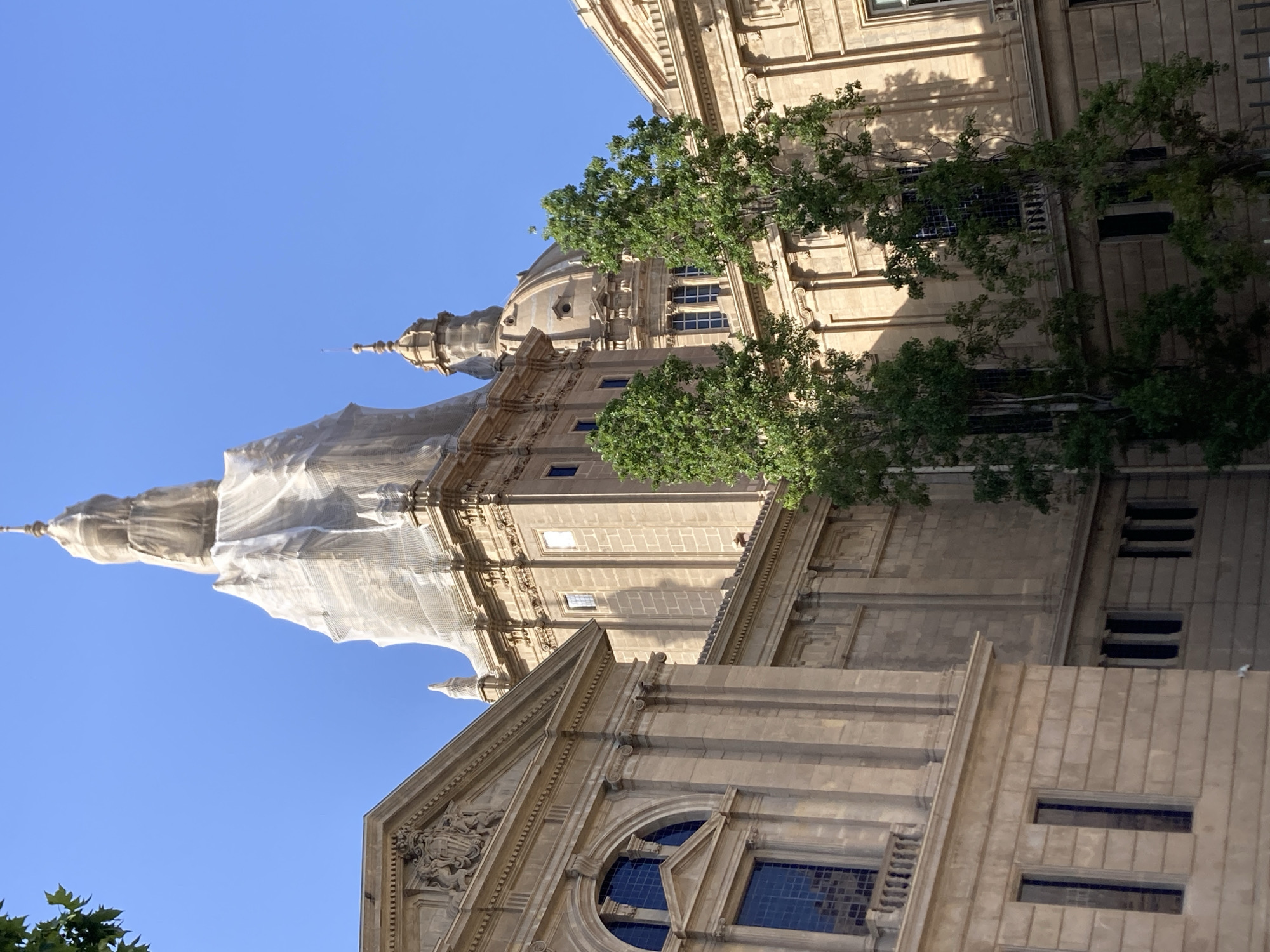 The image shows the Palau Nacional in Barcelona, Spain. The building is a large, neoclassical structure with a central dome and two towers. It is covered in scaffolding and netting, suggesting renovation or maintenance work. Trees are visible in front of the building, and the sky is clear and blue. The architectural style is grand and ornate, with stone facades, arched windows, and decorative elements. The overall impression is one of a historic and significant landmark.