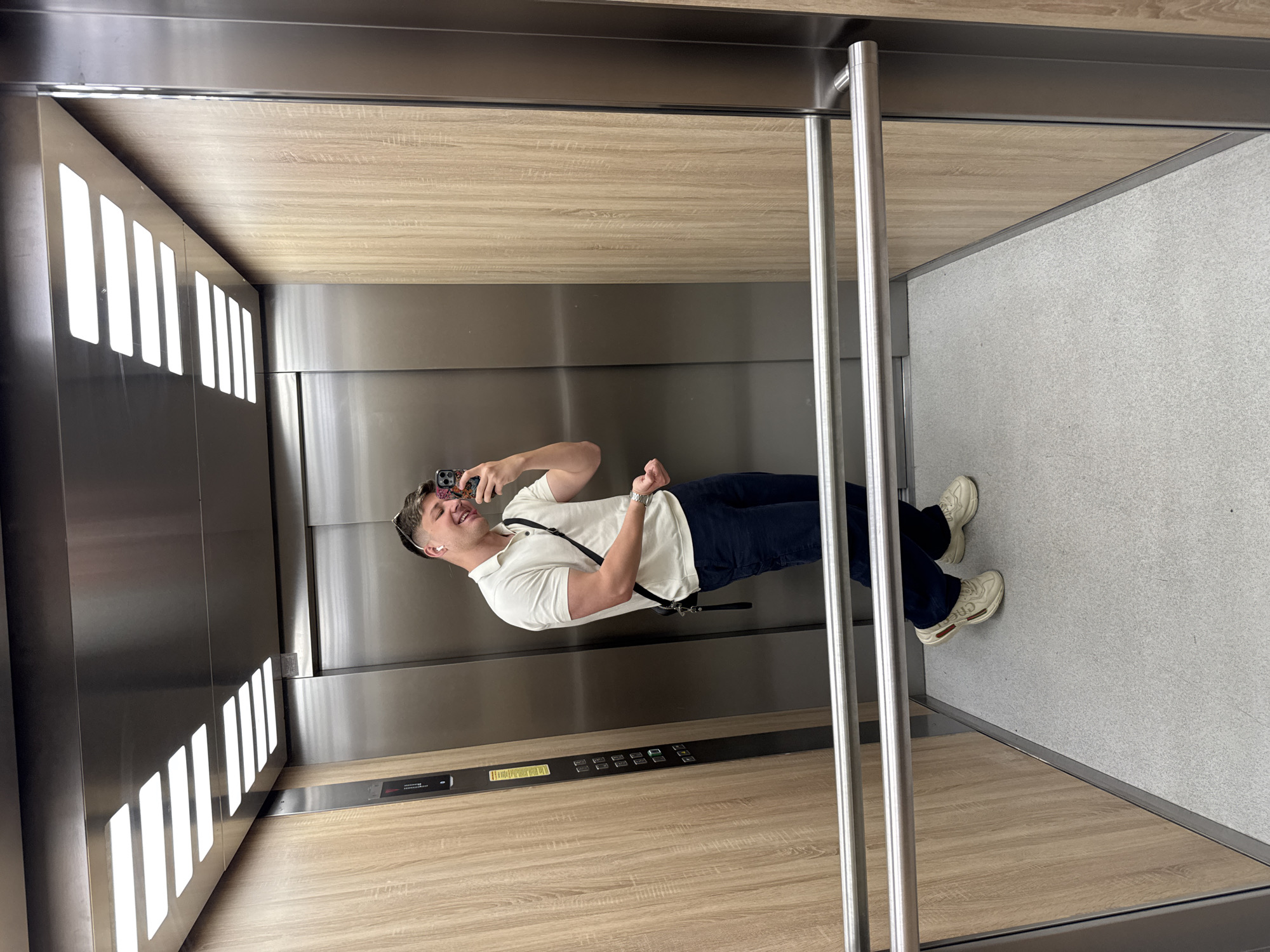 A young man is taking a selfie in an elevator. The elevator's interior consists of wood paneling on the upper walls, a steel panel on the lower walls, a grey carpeted floor, and vertical steel grab bars. The elevator buttons are visible on the steel panel, and there are lights on the left side of the elevator. The man is wearing a white collared shirt, dark blue pants, and light-colored sneakers. He is holding his phone up to take the photo. There is no indication of the location being Plovdiv, Bulgaria from the image.