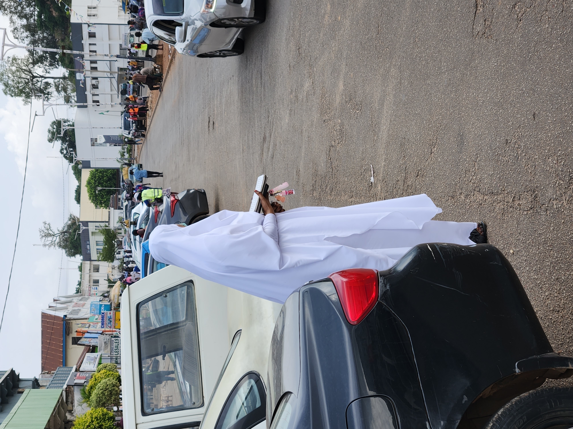 A full-body shot of a person standing on a city street between two vehicles. The person is wearing all white clothing, and is reading from a book. In the person’s left hand are small objects that appear to be ribbons or flowers. The city streets are lined with buildings and parked vehicles. There are other people farther down the street and in front of the buildings.