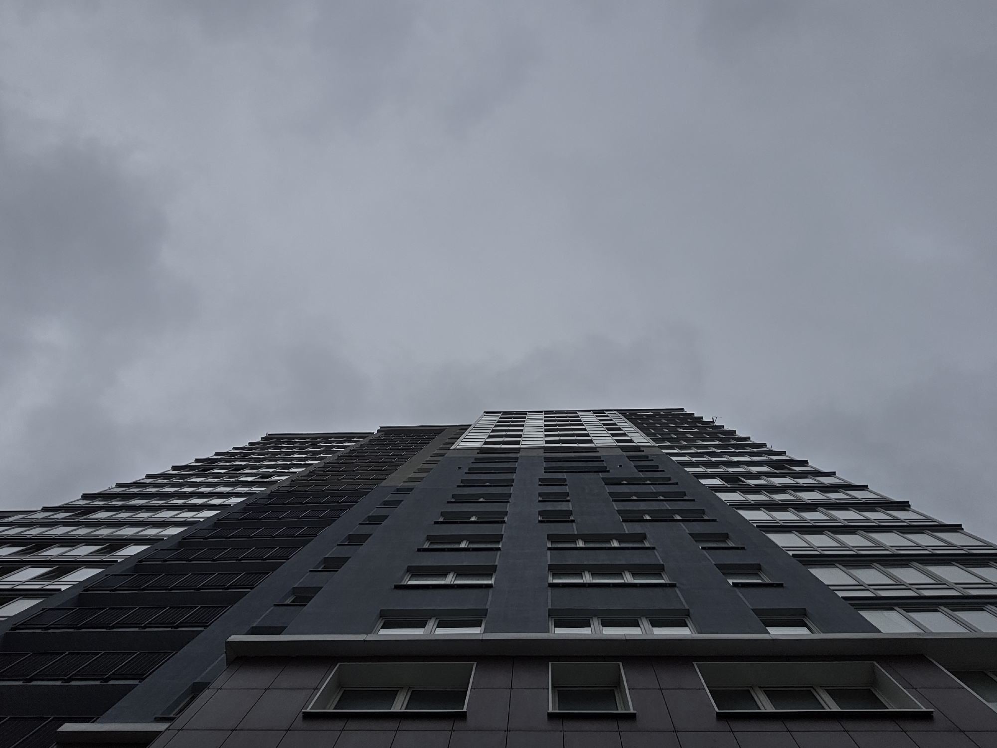 This image captures a modern, multi-story building under an overcast sky, presented from a low-angle perspective. The photograph is oriented such that the building extends horizontally across the frame, originating from the lower-left corner and tapering towards the upper-right, implying a tall structure viewed from below and rotated.
The building's facade is primarily dark grey and charcoal, featuring distinct architectural sections. The closest portion on the left exhibits a dark grey, tiled or panelled exterior with several rectangular, white-framed windows, two of which are visible in the foreground. Adjacent to this, a section with a smooth, darker grey facade is characterized by multiple rows of uniform, vertically elongated, rectangular windows with light frames. Above this, a different segment of the facade presents a series of dark, rectangular louvers or vents interspersed with windows. Further along the building, towards the right, the facade changes again, showcasing a pattern of larger, reflective glass panels or windows. The building continues beyond the frame, suggesting significant height and length.
The sky, occupying the upper-right and central-right portions of the image, is uniformly grey and heavily clouded, indicating an overcast day with diffused lighting. No direct sunlight, distinct sun, or blue sky is visible. The scene is devoid of people, vehicles, or any discernible landscape features. The location is in the city of Unknown, Unknown.