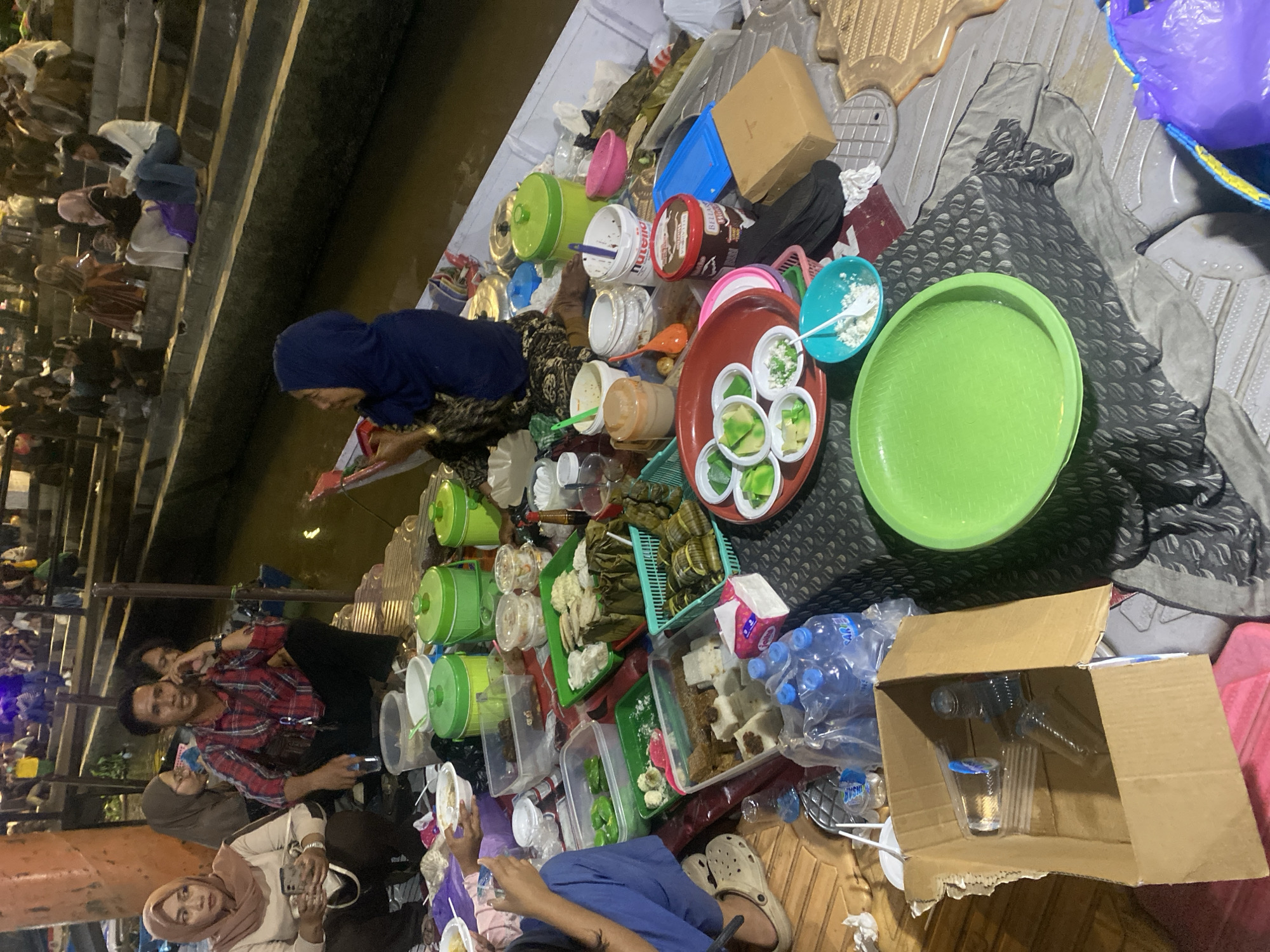 The image depicts a lively outdoor market scene at night in Banjarmasin, Indonesia, likely a floating market due to the presence of water and boats. In the foreground, several individuals are seated on the ground or on low surfaces, surrounded by an assortment of food items displayed in various containers, including plastic tubs, baskets, and banana leaf parcels. People are seen interacting, with some holding phones and others engaged in conversations. A prominent display of colorful dishes, including what appear to be snacks and desserts, are laid out, suggesting a food-selling or communal dining environment. The background shows more people and the tiered structure of the market, with reflections on the water indicating its watery setting. The atmosphere appears to be one of a bustling marketplace with casual activity.