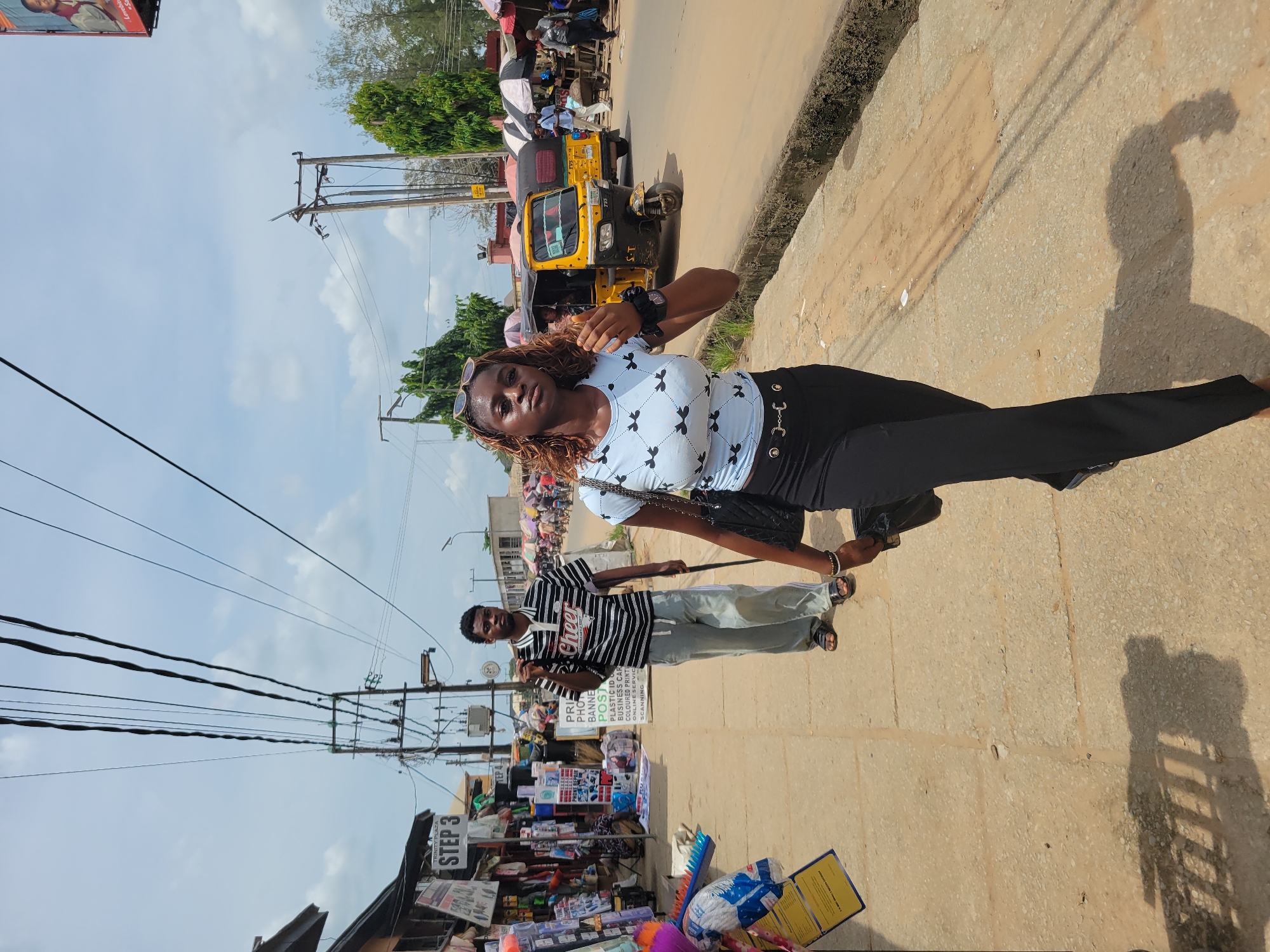A sunlit street scene in Egbagu, Nigeria features a young woman posing for a photo on a sidewalk. She wears a light blue top with black bow patterns, black pants, and sunglasses atop her head. A man in a striped shirt and light-colored pants stands behind her, holding a stick or rod. A yellow "keke napep" (auto-rickshaw) is parked on the road nearby. Various shops and vendors line the street, displaying goods such as stationery, balloons, and printed materials. Power lines stretch across the sky above the street. A sign reading "STEP 3" is visible. The sidewalk appears to be made of concrete slabs. The image is taken at an angle.