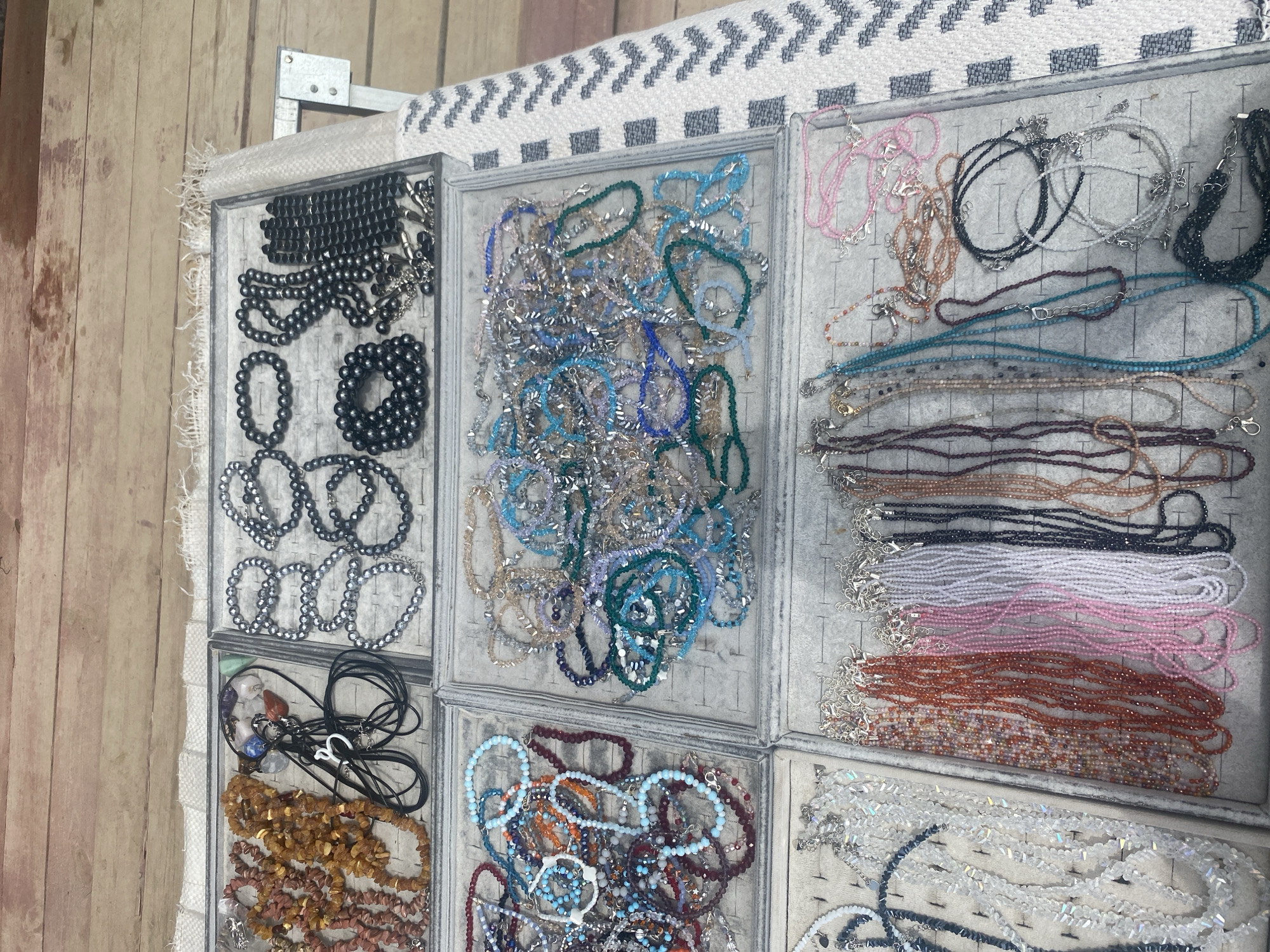 In Arani, Russia, a close-up, overhead shot captures a vibrant display of handcrafted jewelry. The scene, likely at a marketplace or artisan stall, showcases an assortment of beaded necklaces and bracelets meticulously arranged in grey velvet trays. The jewelry features a diverse palette of colors, including earthy tones like amber and brown, deep reds, soft pinks, pristine whites, and various shades of blue and green. Some pieces are simple strings of beads, while others are more elaborate, incorporating metallic accents and intricate designs. The background suggests a rustic setting with weathered wooden planks and a textured textile runner adorned with a geometric pattern. The lighting is even, illuminating the fine details of each bead and clasp, creating a rich visual tapestry of color and texture.