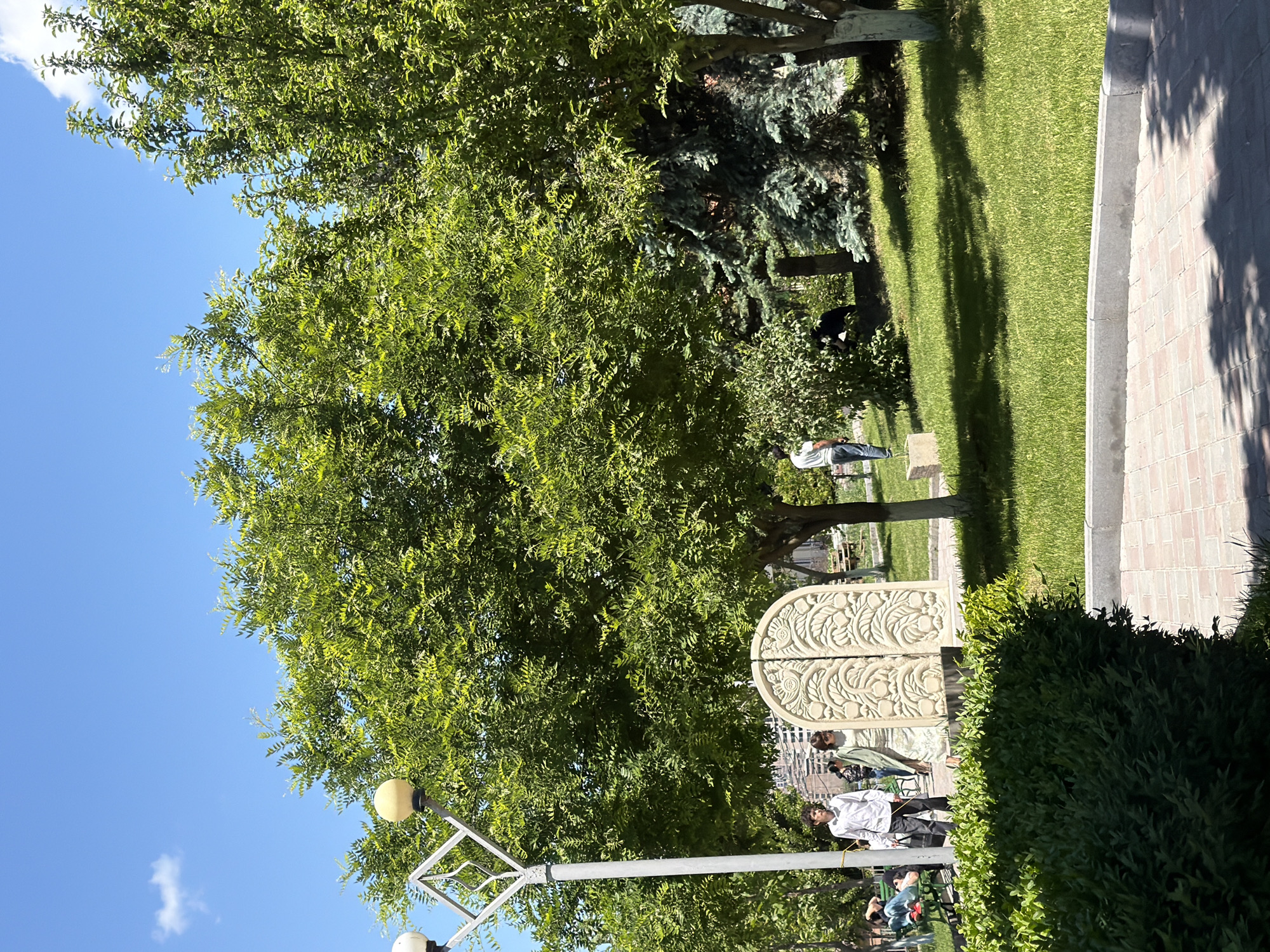 The image depicts a park in Yerevan, Armenia, showing a lush green environment under a clear blue sky. A large tree with dense foliage dominates the upper left of the frame. In the foreground, a stone bench or structure with intricate carvings is partially visible. Several people are present in the background, near benches and pathways, appearing to engage in leisure activities. The park features neatly maintained grass, paving stones, and various trees providing shade. A street lamp is visible in the foreground.