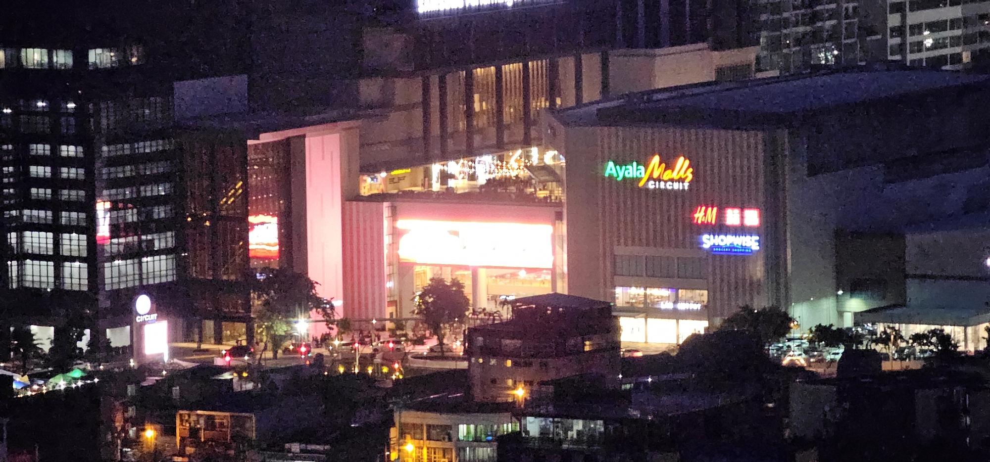 This is a nighttime aerial shot of Ayala Malls Circuit in Makati City, Philippines. The mall features a prominent "Ayala Malls CIRCUIT" sign with multi-colored neon lettering. Below this, "H.M." and "SHOPWISE" signs are also visible. The mall is illuminated with bright lights, including a large pinkish-orange digital display board. Surrounding the mall are other buildings, some with illuminated windows, indicating urban density. Streetlights and vehicle lights suggest active traffic. Trees and dark foreground elements hint at landscaping and lower structures. The scene captures a lively commercial and urban environment at night.