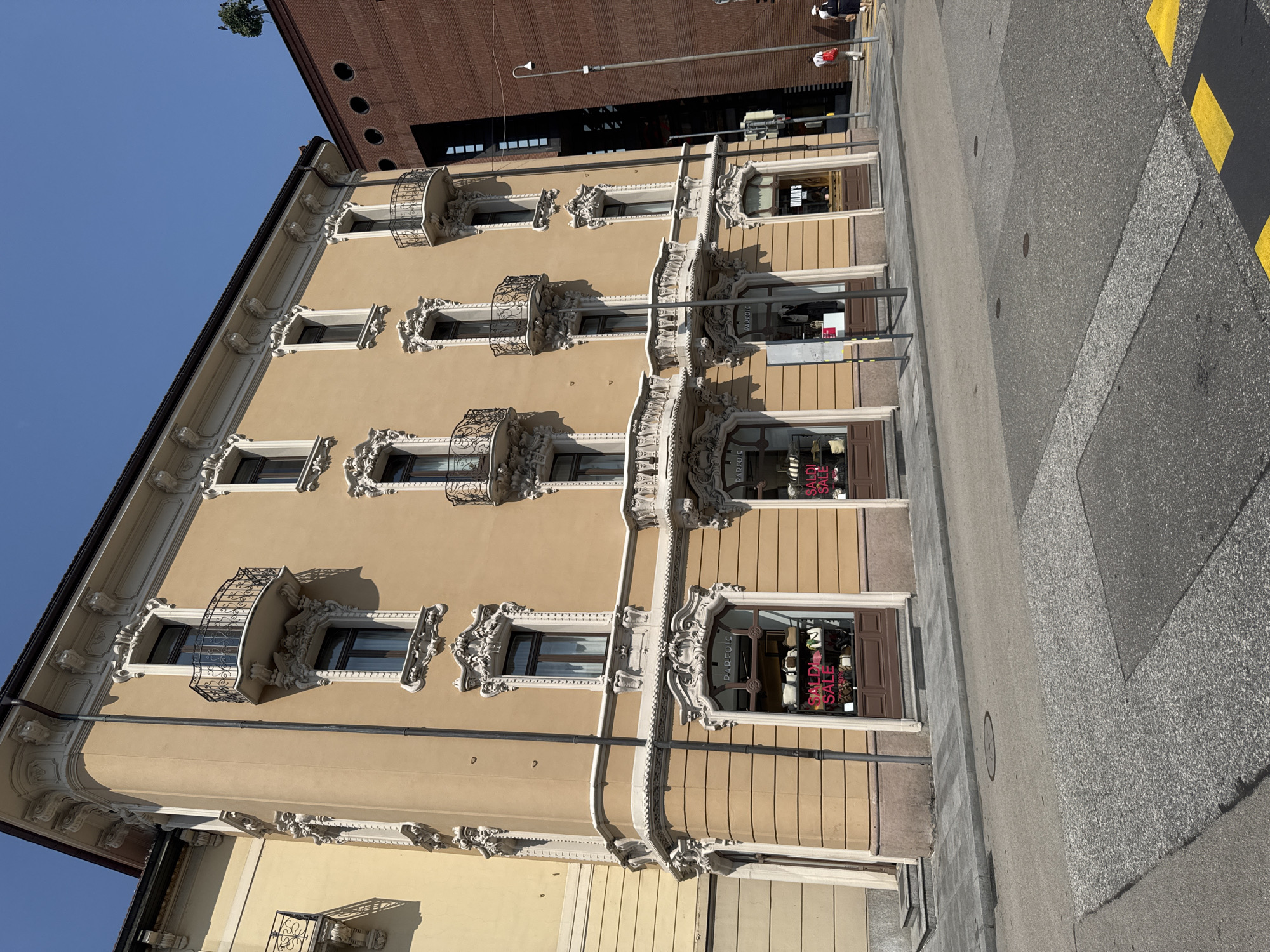 The image depicts a street view in Lugano, Switzerland, showcasing the facade of an ornate, multi-story building with a light yellow stucco finish. The building features intricate white decorative mouldings around its windows and balconies, which are adorned with black wrought-iron railings. Several shops are visible at street level, with signs indicating "PARFOIS" and "HOLIDAYS". Pink "SALDI SALE" signage is prominently displayed in some of the shop windows, suggesting a sale event.
Adjacent to this building, a modern, dark red brick structure with circular window openings rises. The street in the foreground is paved with asphalt and features yellow markings, possibly indicating a pedestrian crossing or traffic regulation. A lamppost is positioned on the sidewalk. Two individuals are walking on the sidewalk in the distance, one carrying a red bag. The sky above is a clear, bright blue. The overall scene suggests a commercial area in a European city during daylight hours.