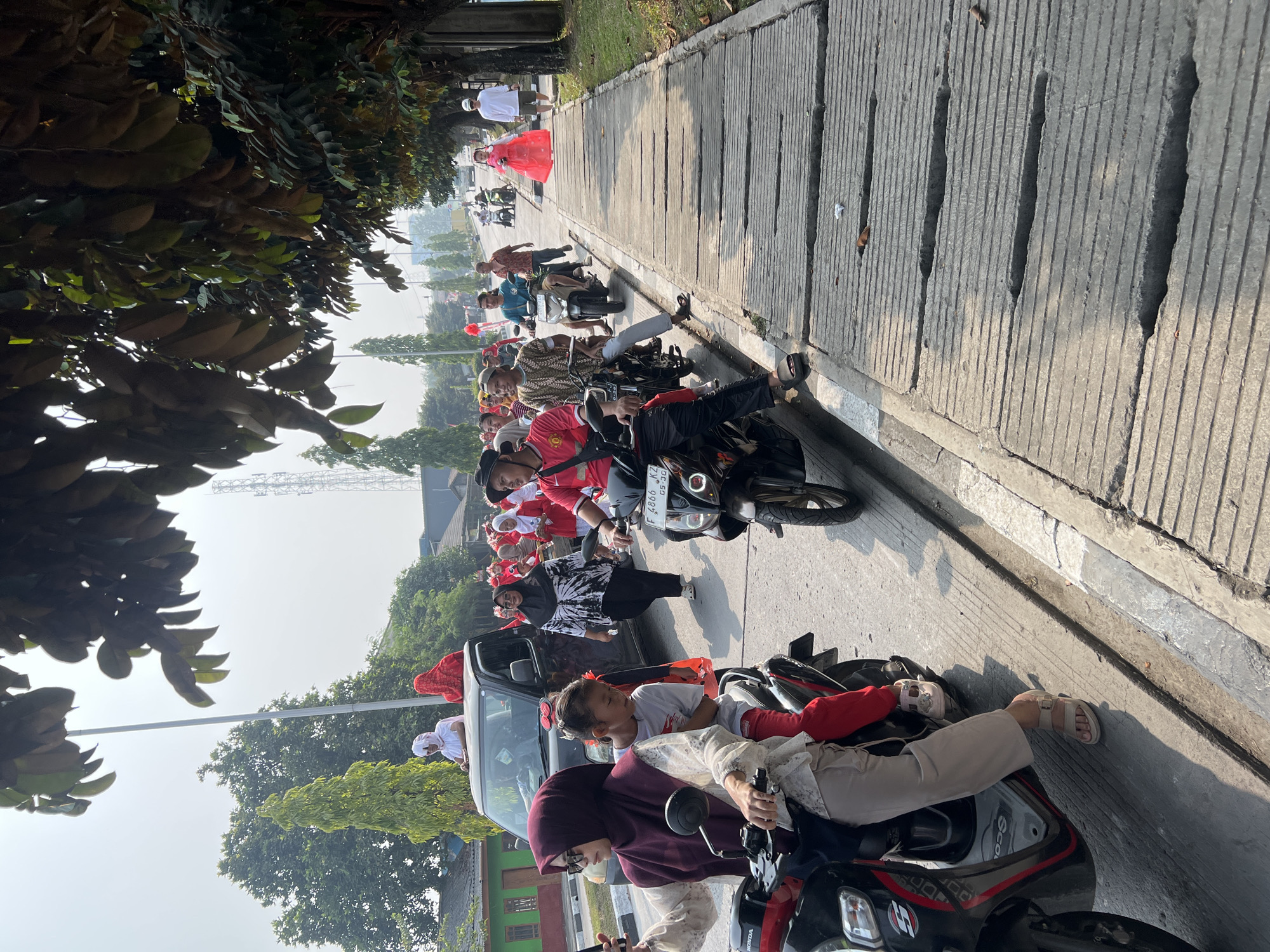A group of people are participating in a procession or parade in Klapanunggal, Indonesia. The scene depicts a street with a sidewalk on the right, lined with trees. Several individuals are on motorcycles, with some carrying passengers. A black minivan is also present, with people standing on top of it. Many participants are wearing red and white attire, suggesting a national celebration or event. One person is walking on the sidewalk in a bright red dress. The overall atmosphere appears festive and active, with a clear sky and bright daylight.