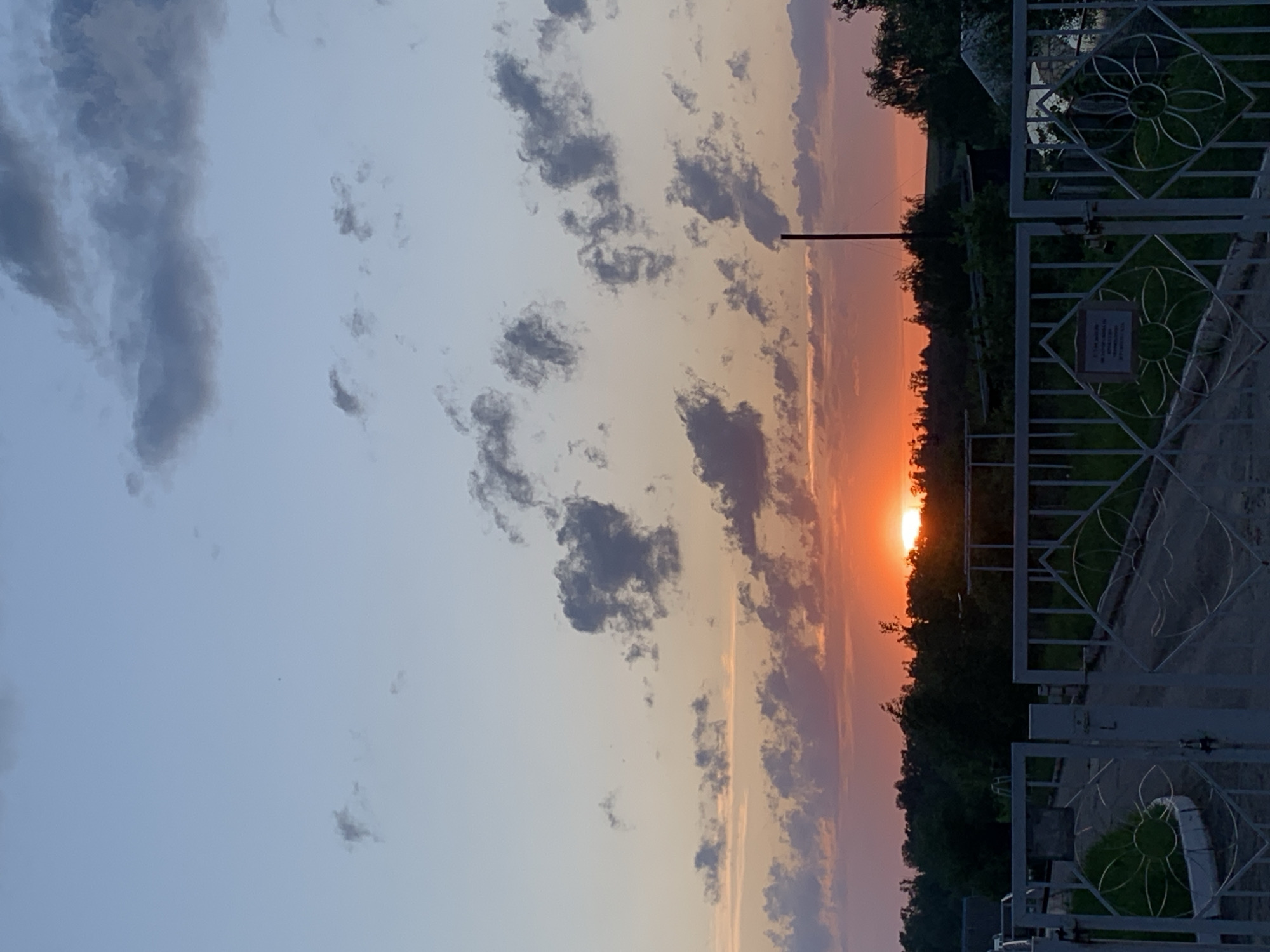 The image shows a sunset in Фоки, Russia. The sun is setting behind a line of trees, casting an orange glow in the sky. There are clouds in the sky, with some of them appearing as dark gray patches against the lighter sky. A metal fence with decorative floral patterns is in the foreground. A pole, possibly a utility pole or light post, is visible against the sunset sky.