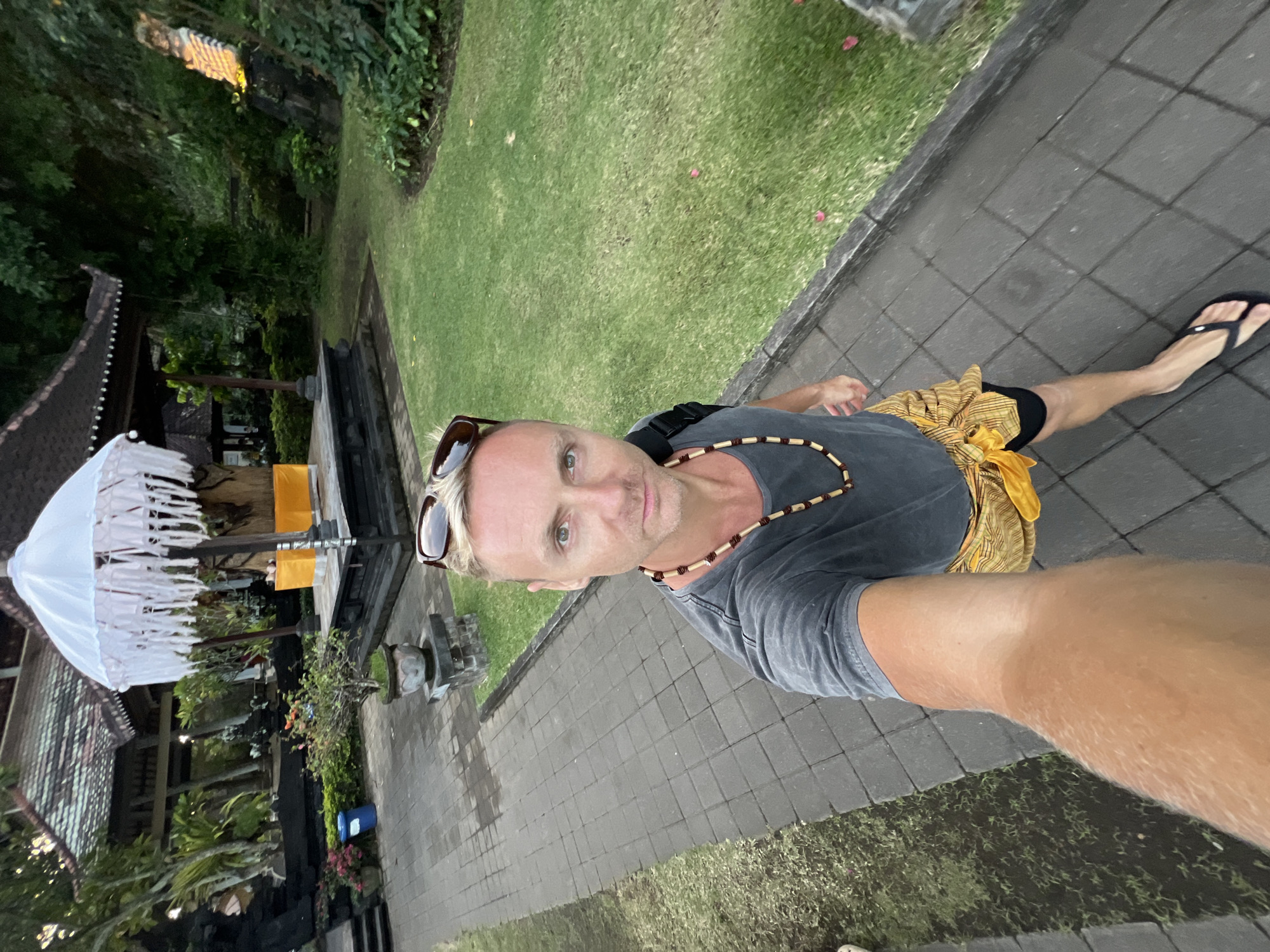A blonde man with sunglasses perched on his head and a beaded necklace around his neck is taking a selfie in a paved outdoor area. He is wearing a dark grey t-shirt, a yellow sarong tied around his waist, black shorts, and black flip-flops. The man is standing on a paved walkway lined with a grassy area and manicured bushes. In the background, there is a traditional Indonesian temple structure with a large white umbrella adorned with white fringe. The temple features dark wood, a tiled roof, and a raised platform with yellow fabric. The environment appears lush and green, with trees and foliage visible. The scene suggests the man is visiting a cultural site in Dawan, Indonesia.