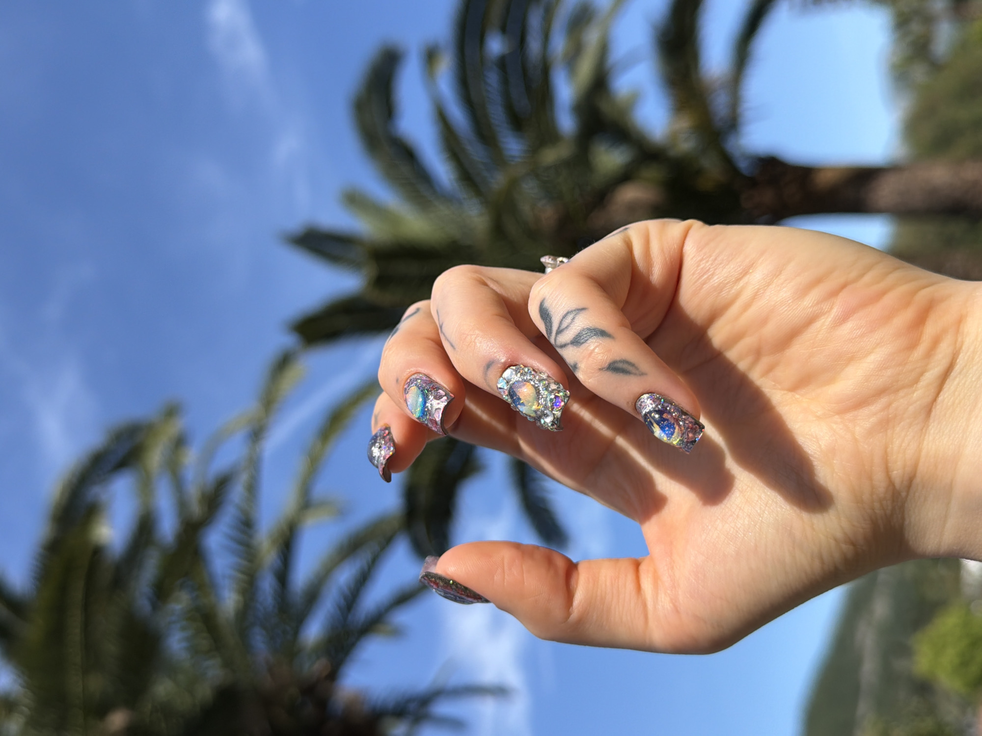 A human hand, likely female, is prominently featured in a close-up against a vibrant blue sky with sparse, wispy white clouds. The hand showcases long, elaborately decorated nails, each adorned with iridescent, chunky glitter and various sparkling, crystal-like embellishments that catch the light. Several small, faded black tattoos, appearing as delicate leaf or abstract patterns, are visible on the fingers. A silver ring with a clear stone is worn on one finger.
In the blurred background, the lush green fronds of several large palm trees are visible, indicating a warm, Mediterranean or tropical setting. The bright, natural sunlight suggests it is daytime, likely midday or early afternoon, with clear and sunny weather. The overall scene evokes a relaxed and pleasant atmosphere, consistent with the provided location context of Budva, Montenegro. There is no visible text in the image.