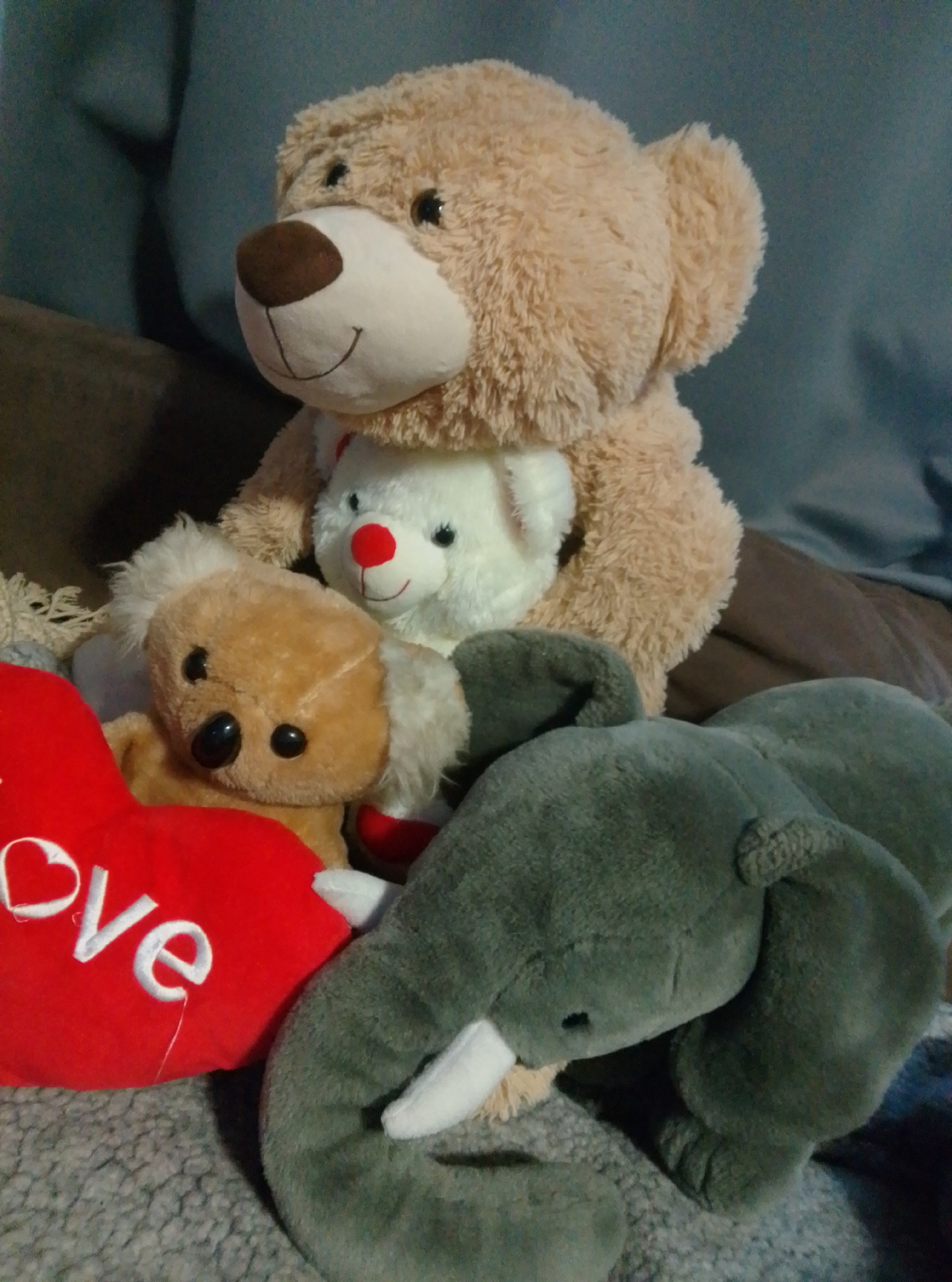 A collection of stuffed animals is arranged on a soft surface. The largest is a light brown teddy bear, positioned at the back and slightly to the left, with its head tilted forward. In front of it, a smaller white teddy bear with a red nose and ears is partially visible. To the left of the white bear, a light brown koala bear is looking forward, with a red heart-shaped pillow with the word "love" embroidered in white in front of it. A large gray elephant stuffed animal is in the foreground, partially obscuring the koala and the heart pillow, with its trunk curled to the left. The background appears to be a couch or soft seating area with a textured gray blanket or rug. The setting is indoors, with no specific landmarks indicating the city of Wedel, Germany.