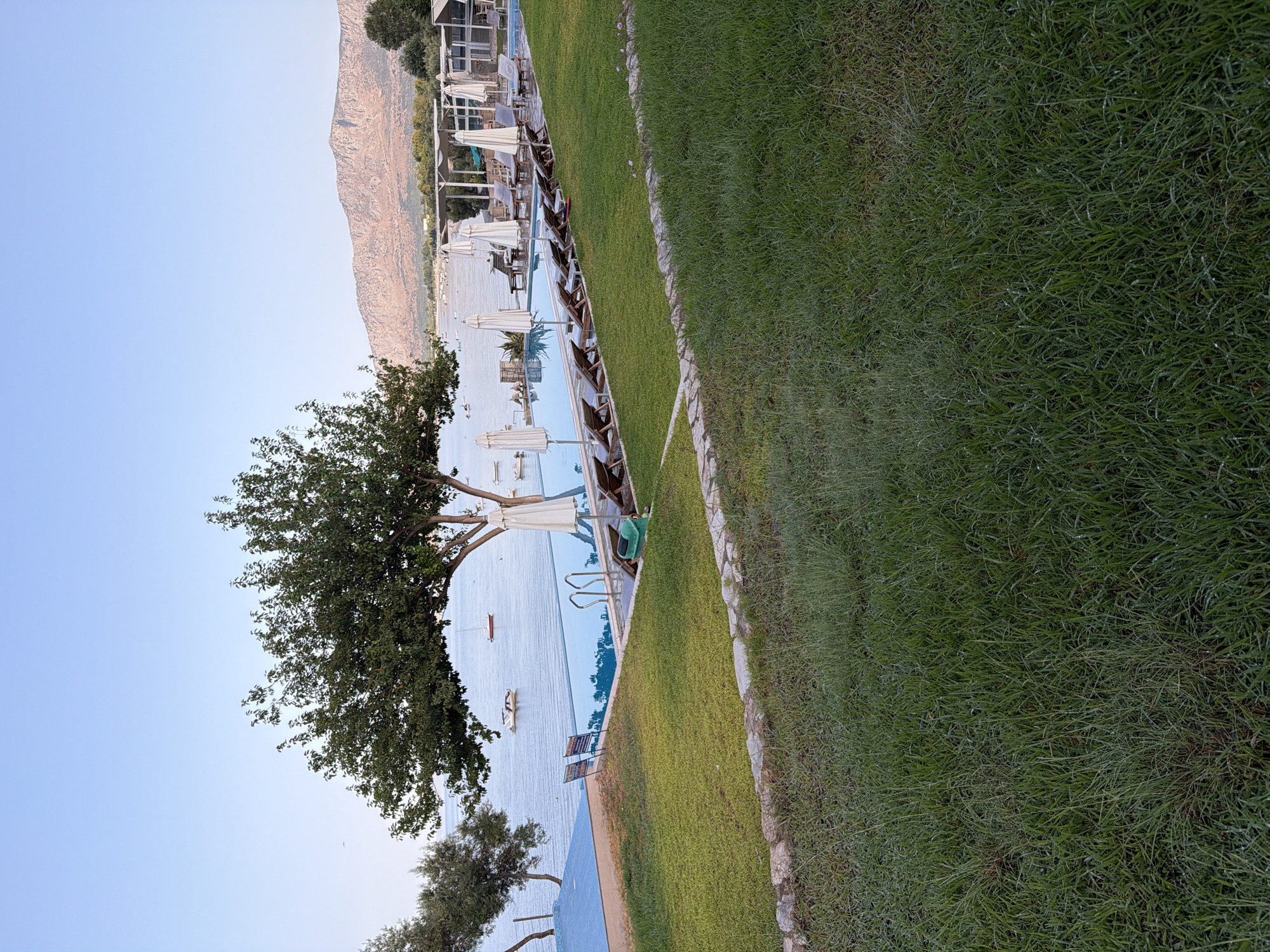 The image shows a resort or hotel pool area next to the sea in the Municipal Unit of Alyzia, Greece. In the foreground, a well-maintained green lawn slopes gently towards a stone border. Beyond the lawn, a swimming pool with clear blue water is visible. Several lounge chairs with white umbrellas are arranged around the pool. The sea is calm and reflects the clear blue sky. In the distance, mountains form the horizon. There are no people visible in the image. The scene suggests a serene and relaxing atmosphere.