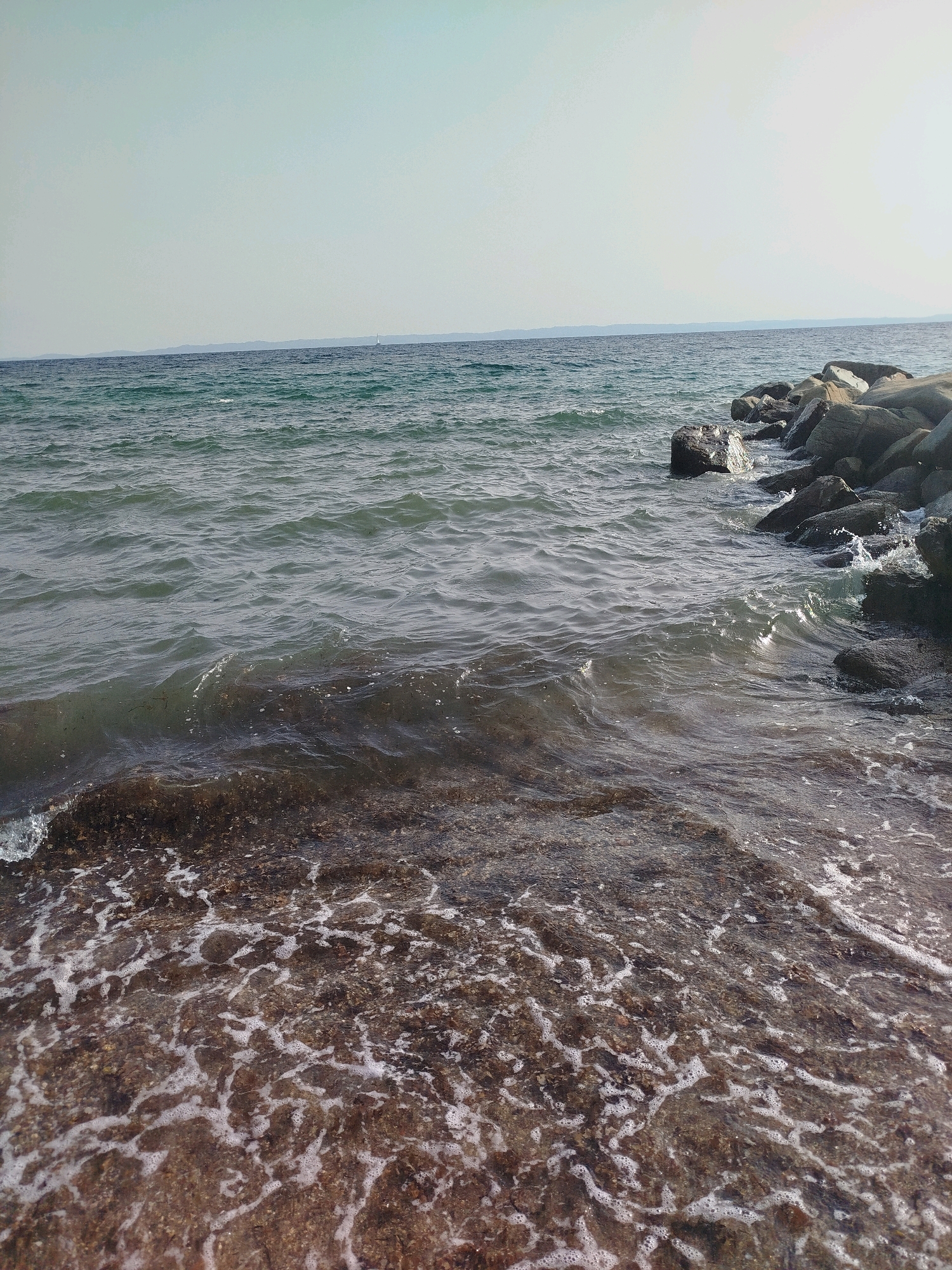 The image depicts a coastal scene in the Sithonia Municipal Unit, Greece. The foreground shows the shallow, murky water of the sea lapping against a rocky seabed covered in seaweed. Small waves create white foam as they break. In the mid-ground, the sea stretches out, appearing dark and somewhat choppy. A line of large, grey rocks forms a breakwater on the right side of the image, with waves crashing against them. In the far distance, a faint, hazy landmass is visible on the horizon. The sky above is a pale, washed-out blue, with a hint of a hazy sun. There are no people or any man-made structures other than the rocky breakwater. The overall impression is one of a natural, rugged coastline on a bright, clear day.
