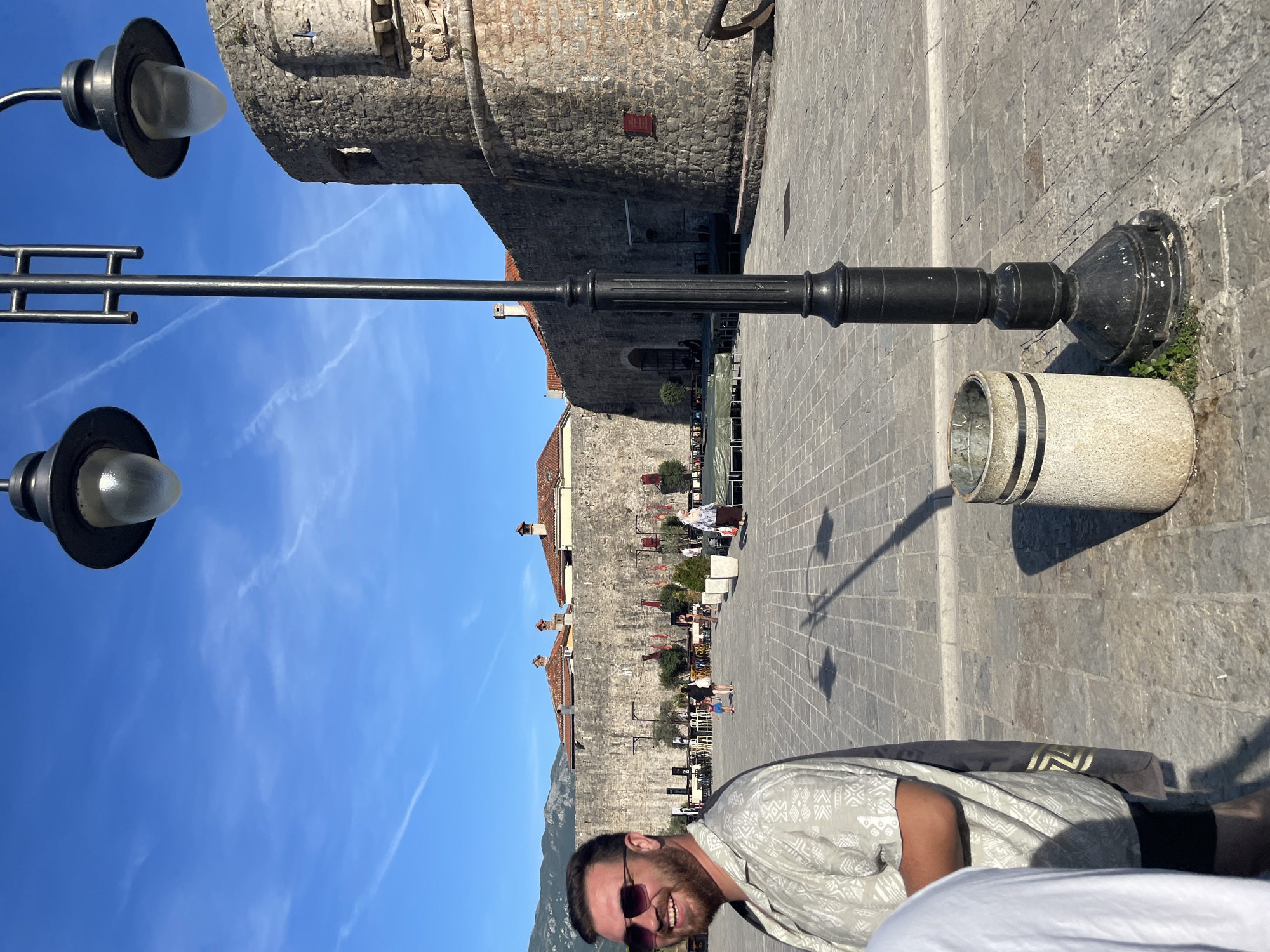 The image shows a sunny day in Budva, Montenegro, with a view of the Budva Citadel walls and towers. In the foreground, a man with sunglasses and a patterned shirt is visible on the left, partially cropped. A tall lamppost with two lights stands in the center of the frame. The ground is paved with cobblestones. In the background, the ancient stone walls of the citadel are prominent, with red-tiled roofs of buildings visible behind them. People are seen walking and sitting in the area in front of the citadel, suggesting it is a public square or promenade. The sky is clear and blue with some wispy clouds.