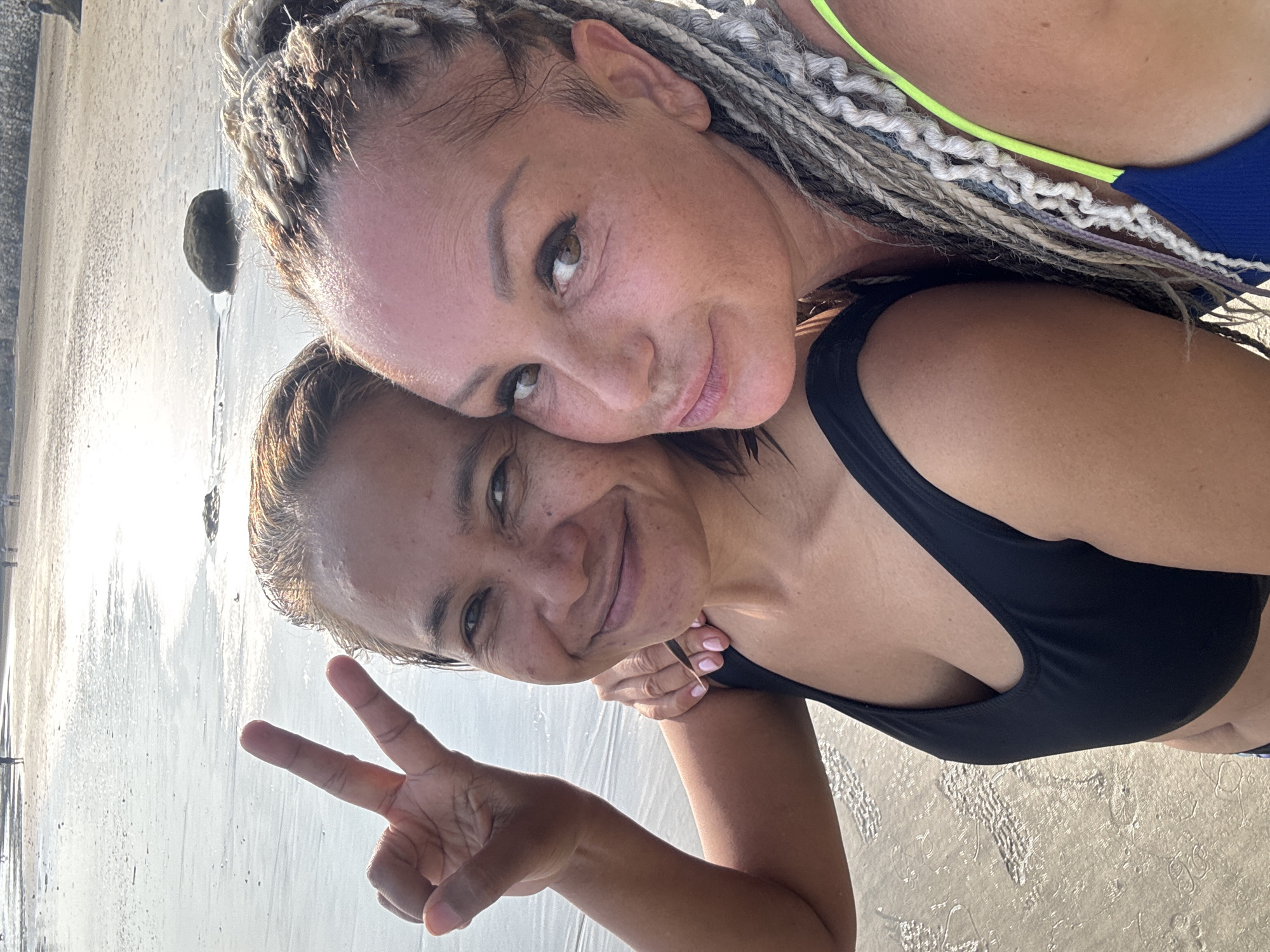 Two women are posing for a selfie on a beach. One woman has braided hair and is wearing a swimsuit with neon green straps. The other woman has dark hair and is wearing a black swimsuit. The woman in the black swimsuit is holding up a peace sign. They are standing on a sandy beach with wet sand and some rocks visible in the background. The location is Jimbaran, Indonesia.